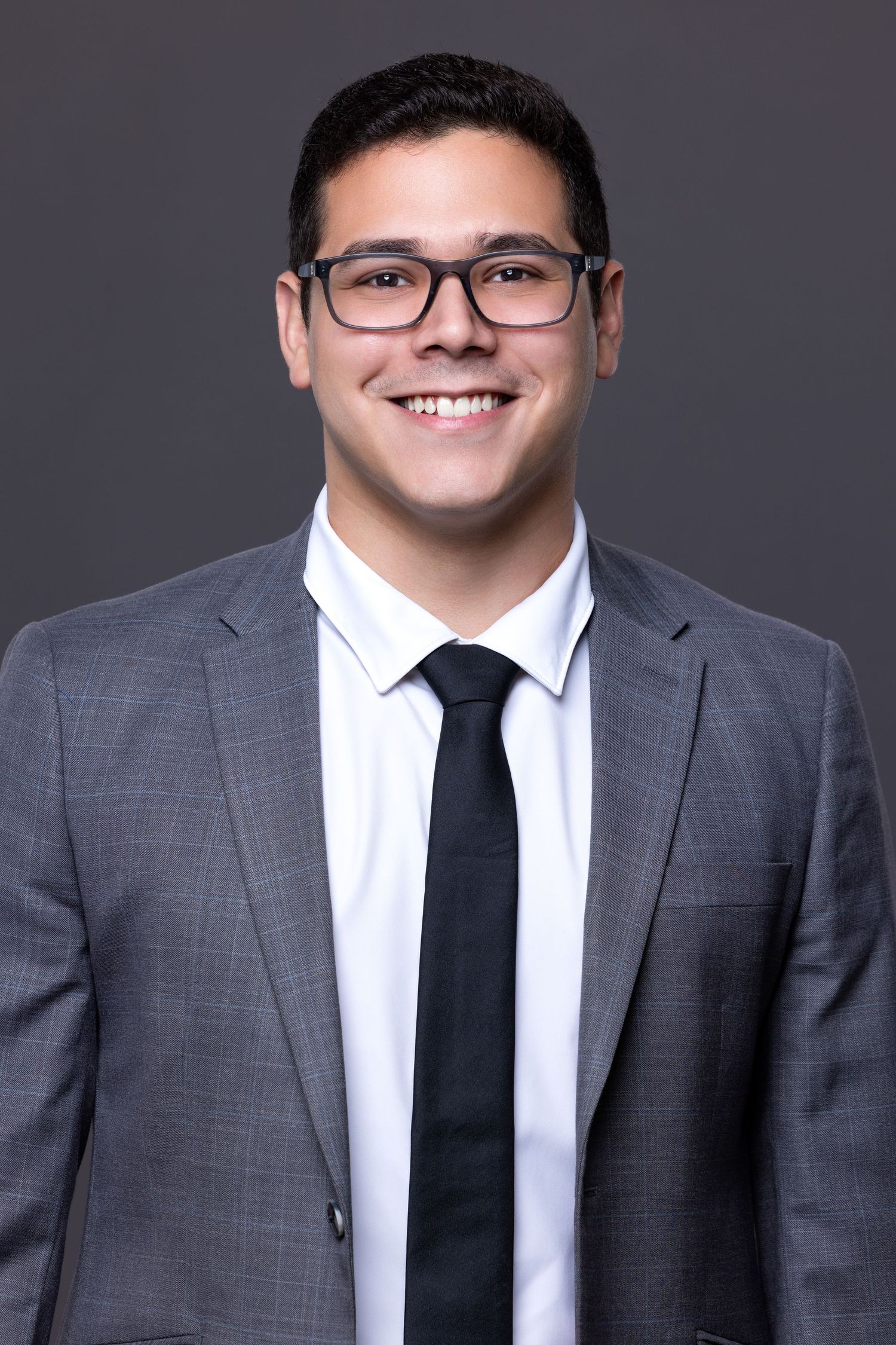 Professional business headshot of person wearing glasses, grey suit and black tie against dark background.