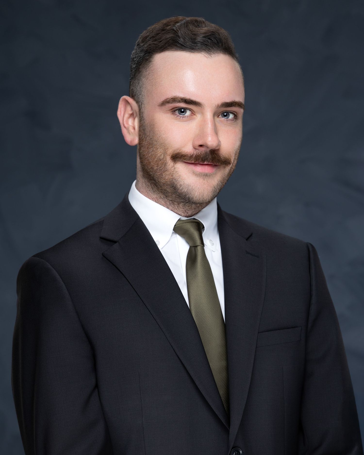 Professional headshot portrait of someone in a black suit and olive green tie against a dark background.
