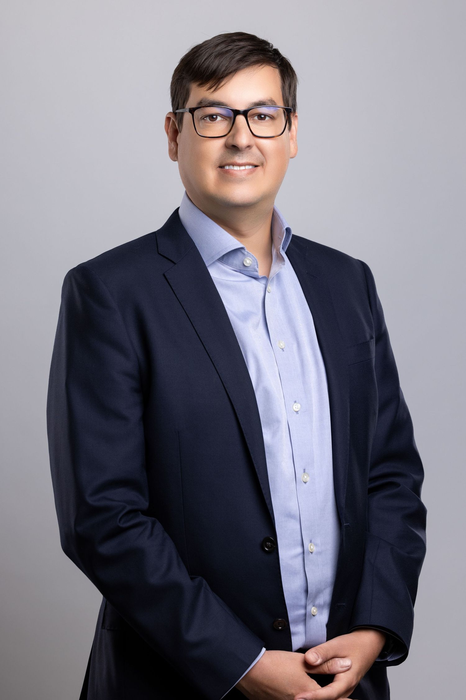 Professional business headshot portrait of a person in a navy suit and light blue shirt against a gray background.