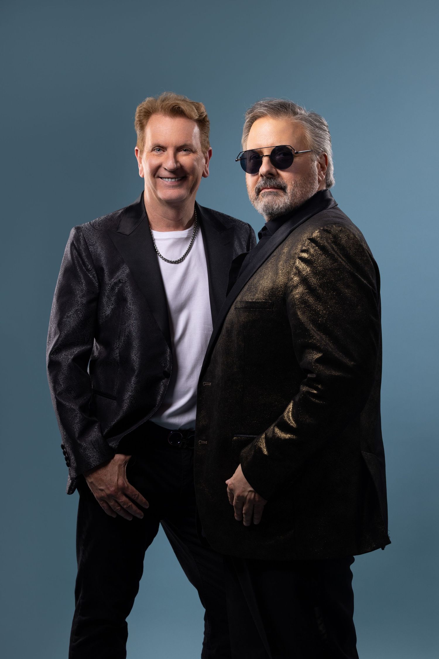Two men in contrasting outfits pose together against a blue backdrop in a series of candid studio shots.