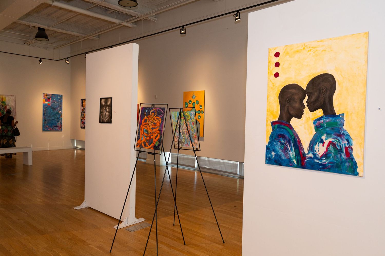 Art gallery interior with white walls displaying colorful paintings and artworks mounted on easels and walls.