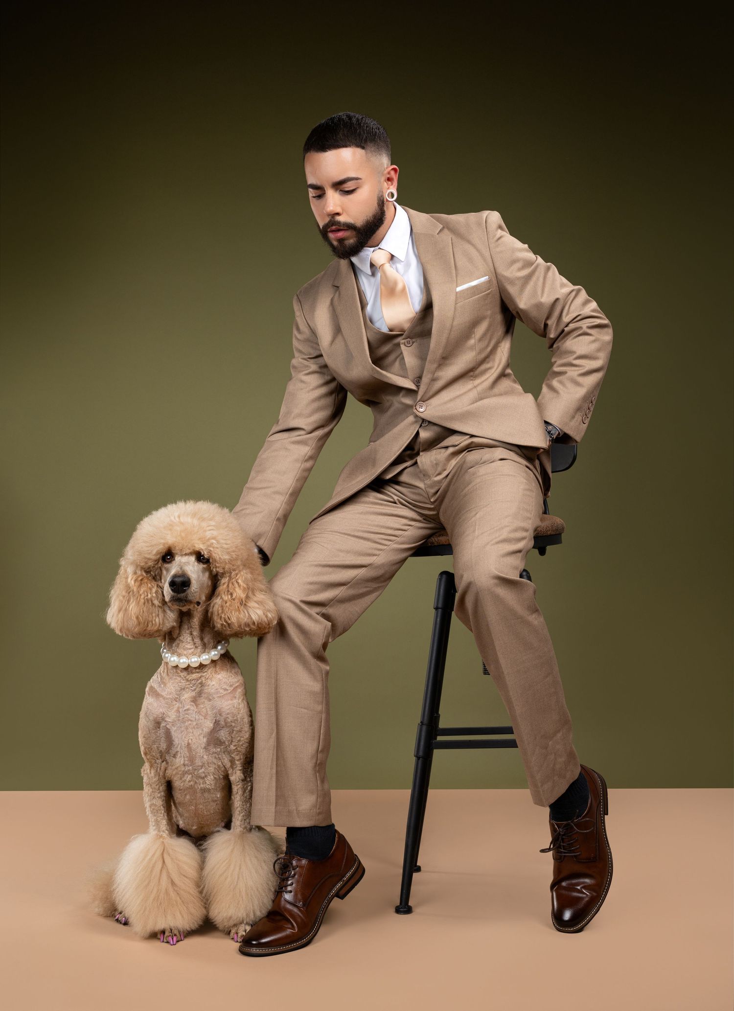 A person in a tan suit poses playfully with a standard poodle in a studio setting against an olive background.