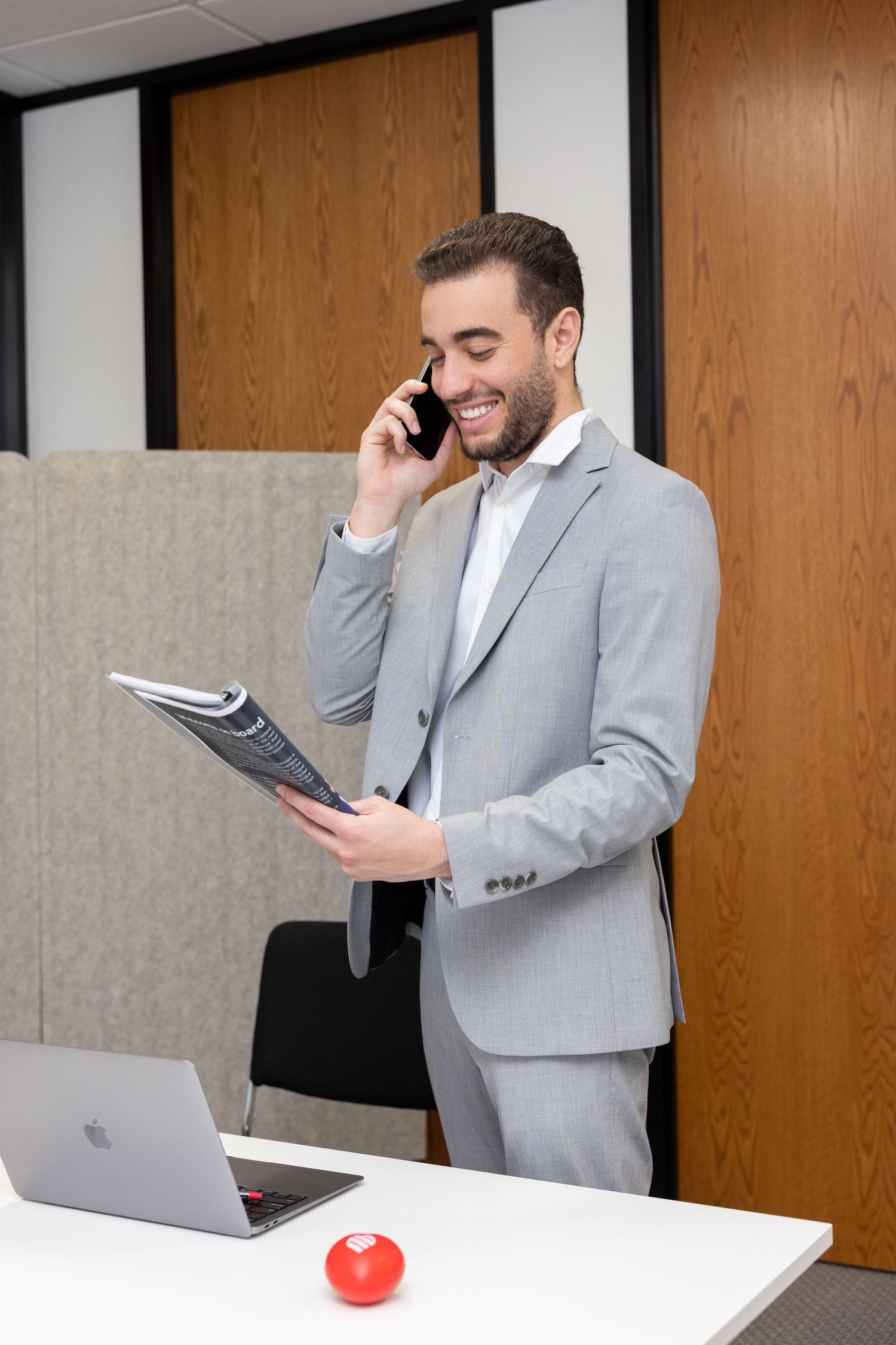 Smiling businessman in gray suit talking on phone while reviewing documents in modern office.