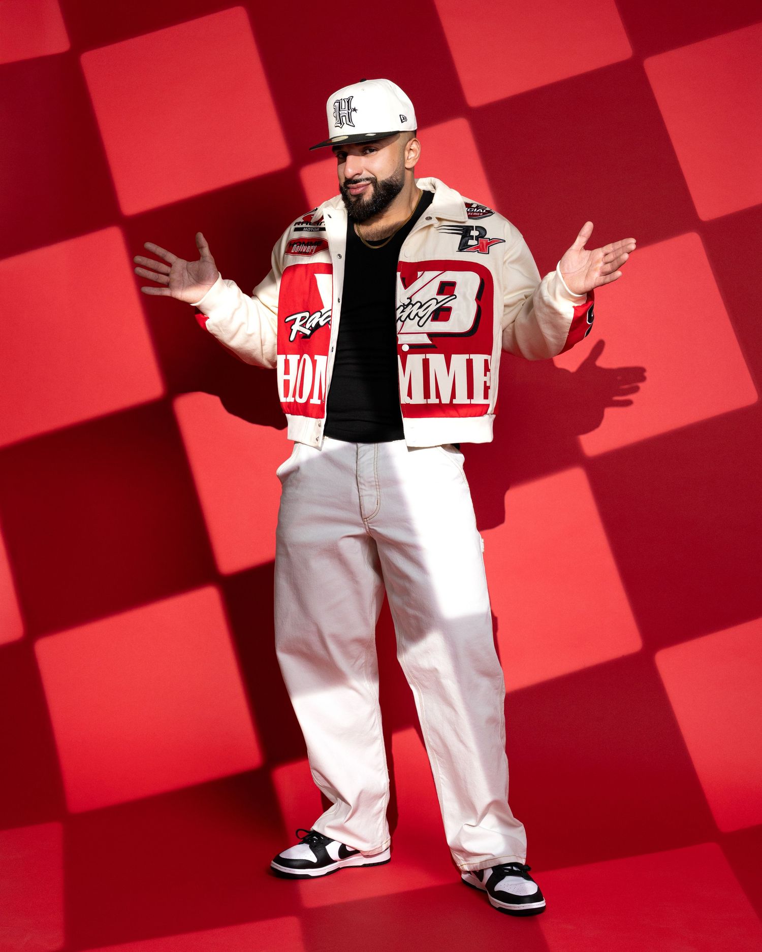 Person in white varsity jacket and baseball cap poses with outstretched arms against red checkered background.