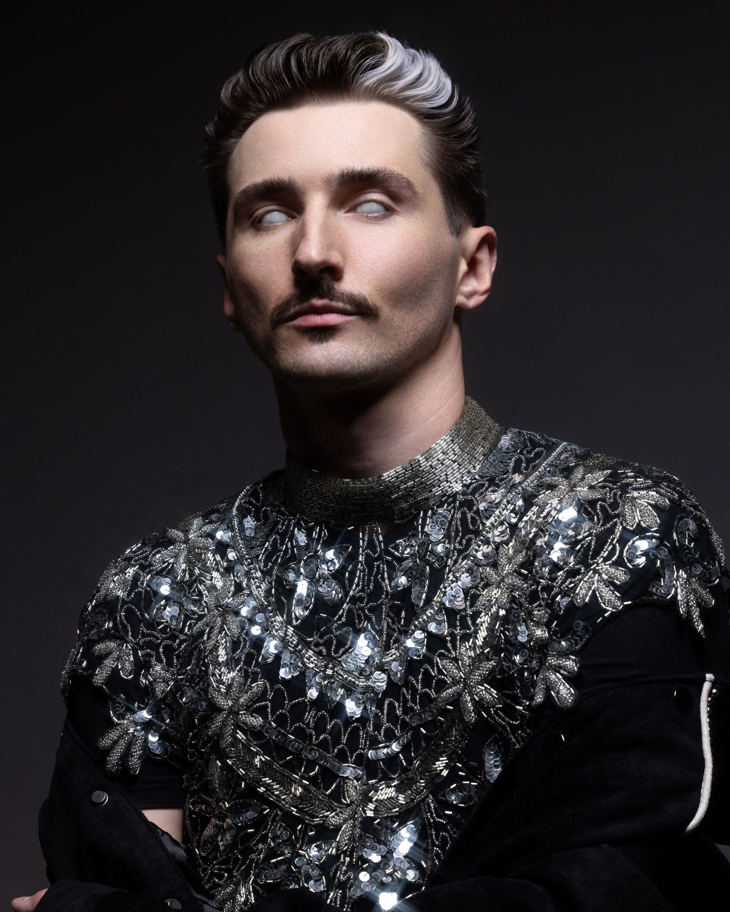 Dramatic portrait of someone wearing an ornate black jacket with crystal embellishments against a dark background.