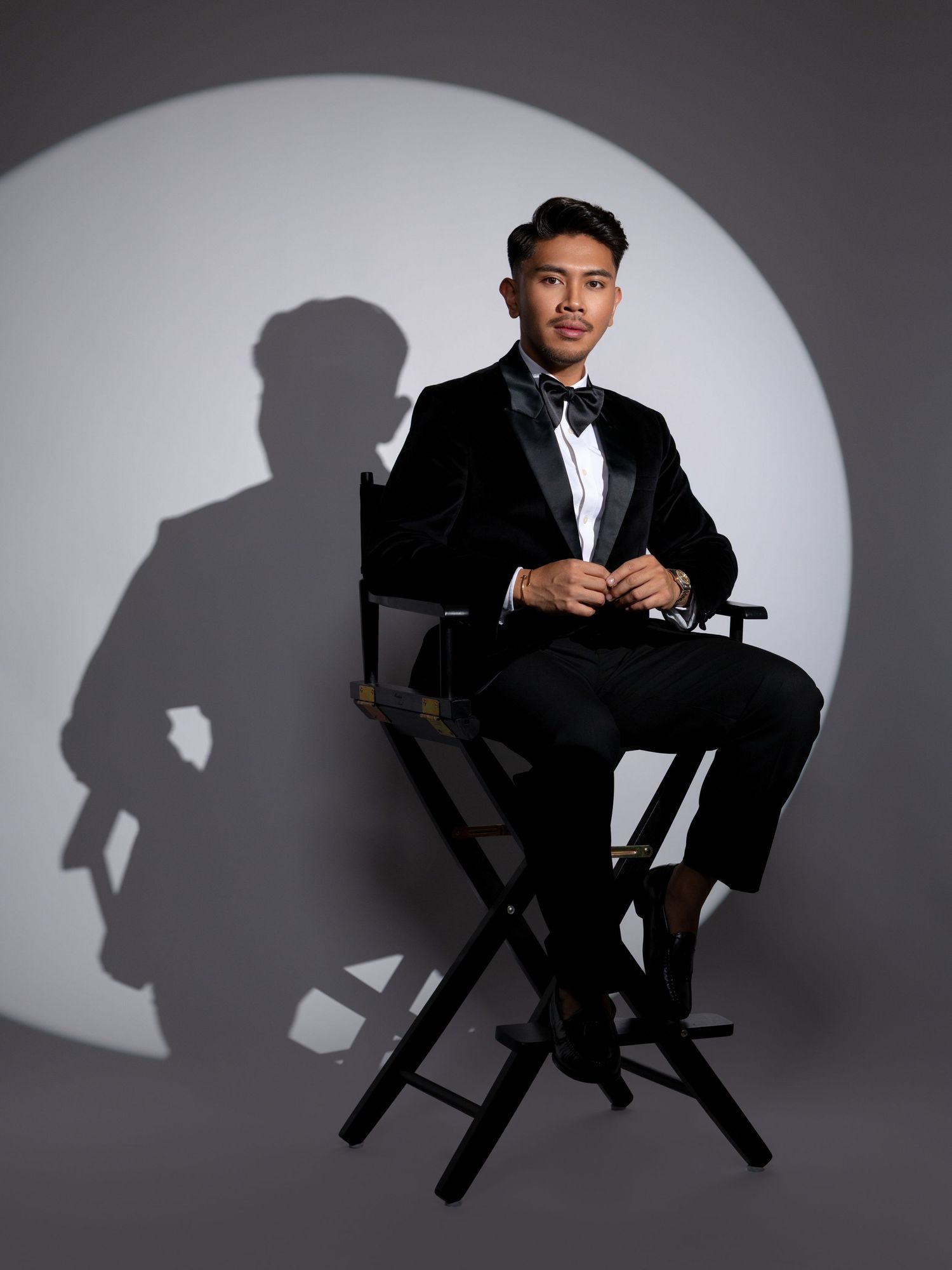 Young man in black tuxedo and bow tie seated in director's chair against dramatic spotlight background.
