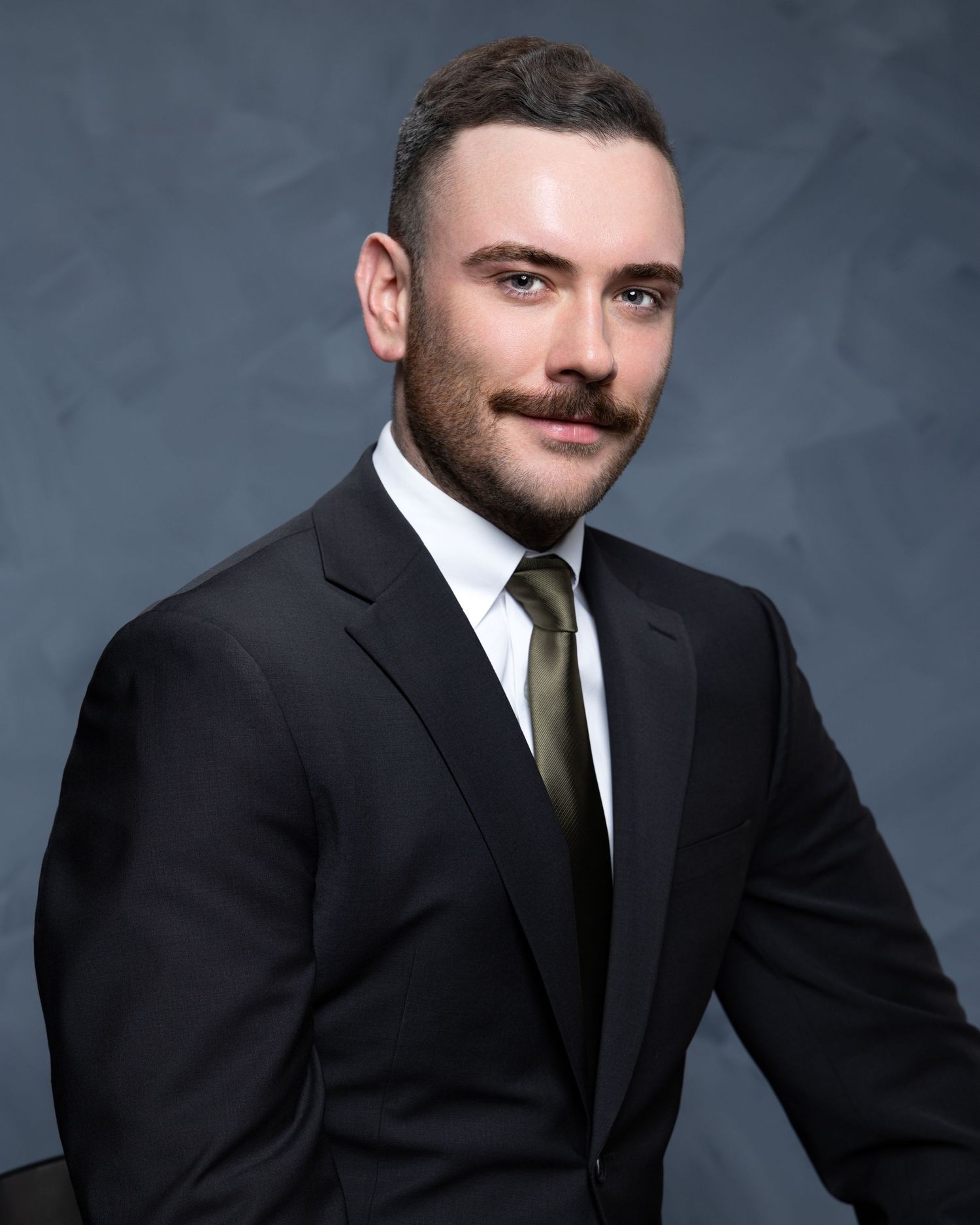 Professional man in black suit with olive tie and mustache poses for formal business headshot photo.
