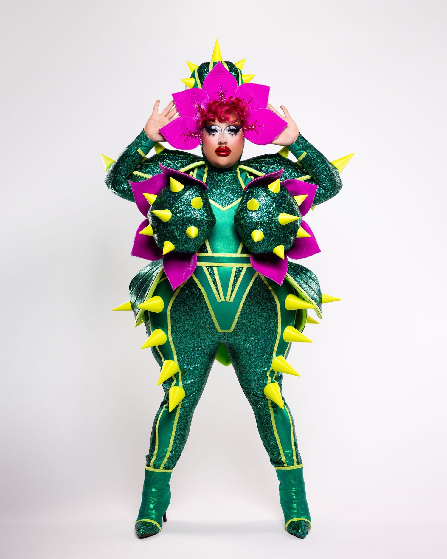 Drag performer in a dramatic emerald green spiked bodysuit with pink hair and yellow accents striking a pose against white backdrop.