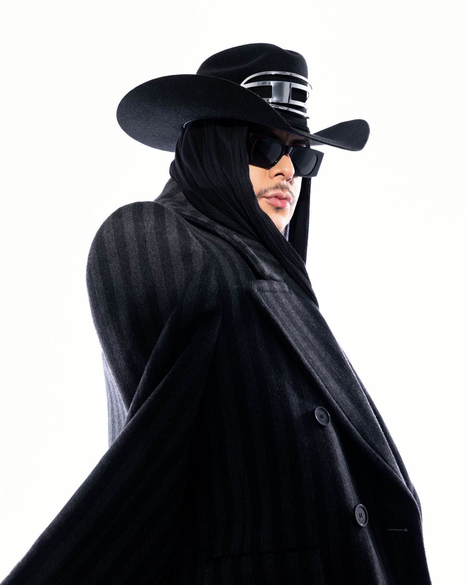 Person in dramatic black wide-brim hat, sunglasses, dark veil, and striped cape coat posing against white background.