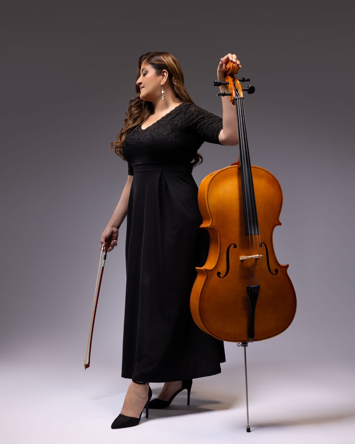 Musician in a black dress holding a cello bow stands elegantly next to a wooden cello against a gray background.