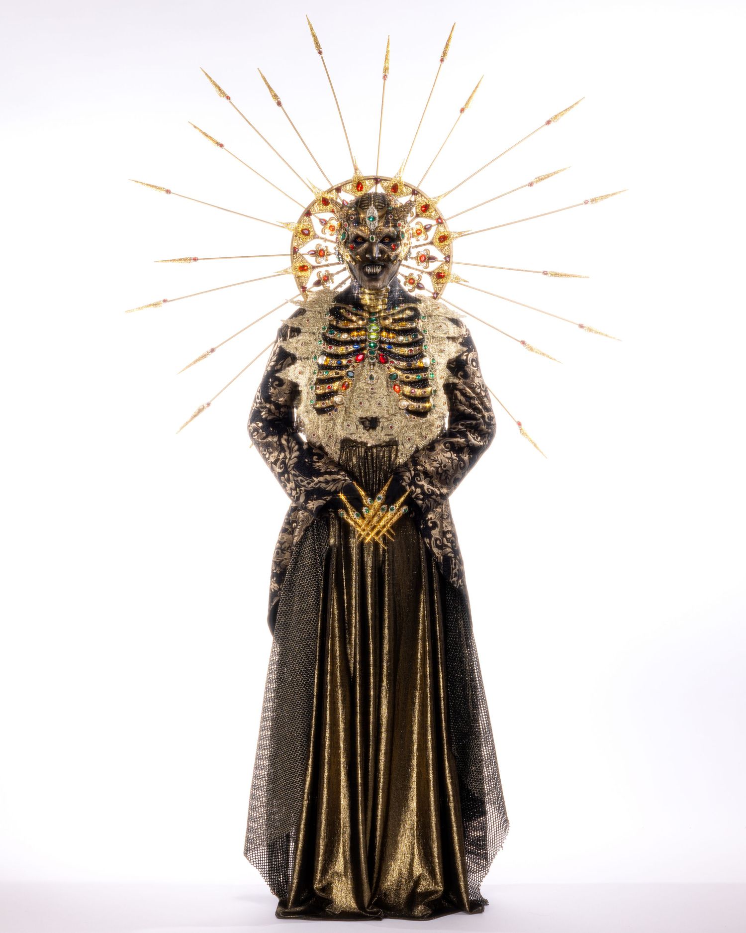 A religious art figure in black and gold robes with a skeleton motif and radiating halo crown against white background.