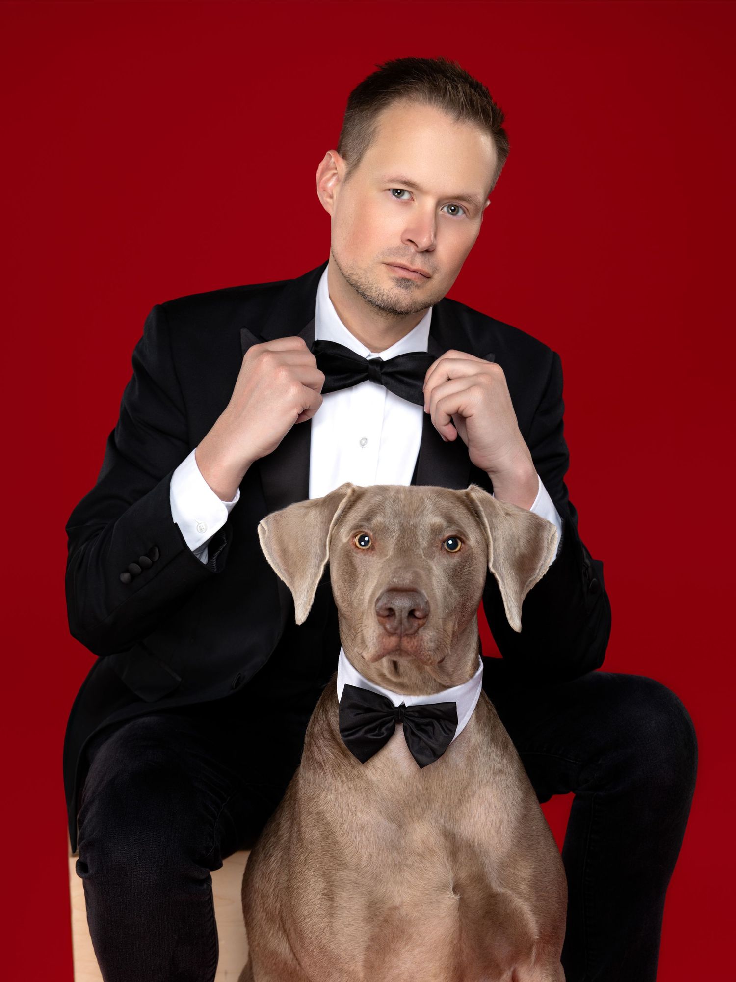 A Weimaraner dog and someone in formal black tuxedos with matching bow ties pose against a red backdrop for a portrait.