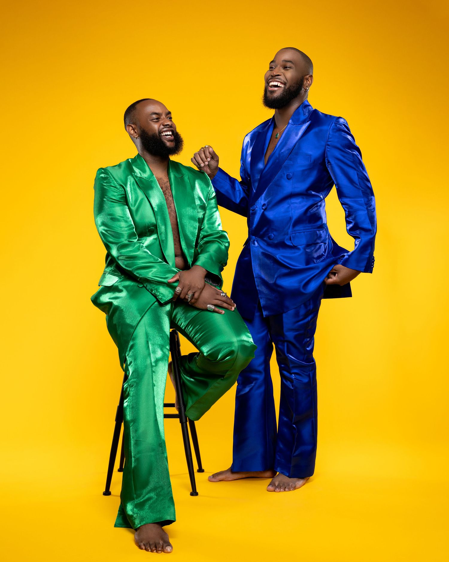 Two people in contrasting silk robes, one blue and one green, share a playful moment against a bright yellow background.