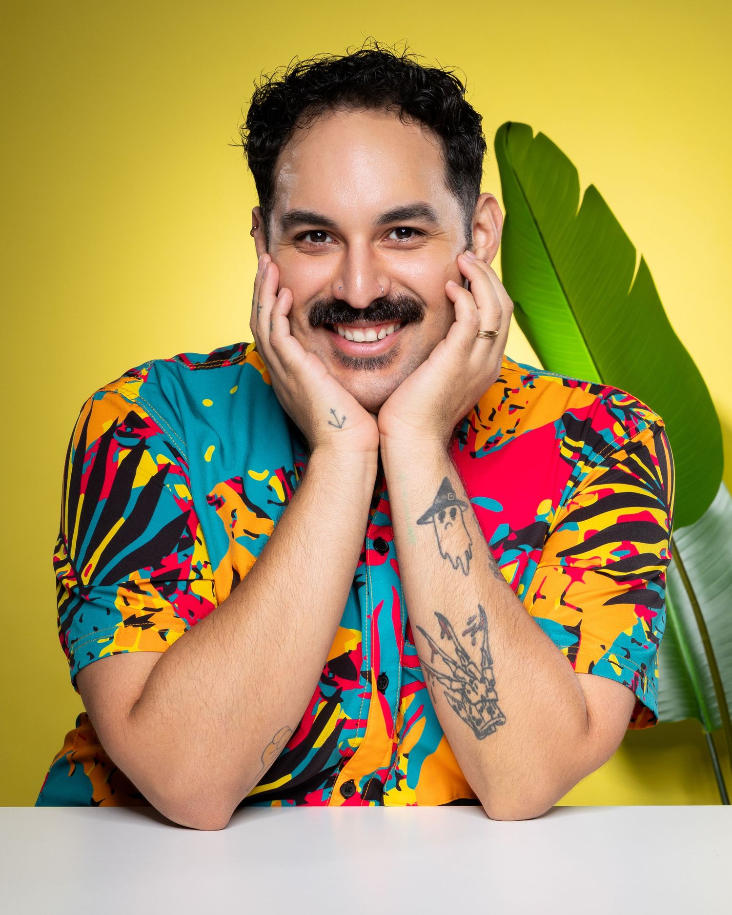 Person wearing vibrant multicolored Hawaiian-style shirt poses playfully against yellow background with tropical plant.