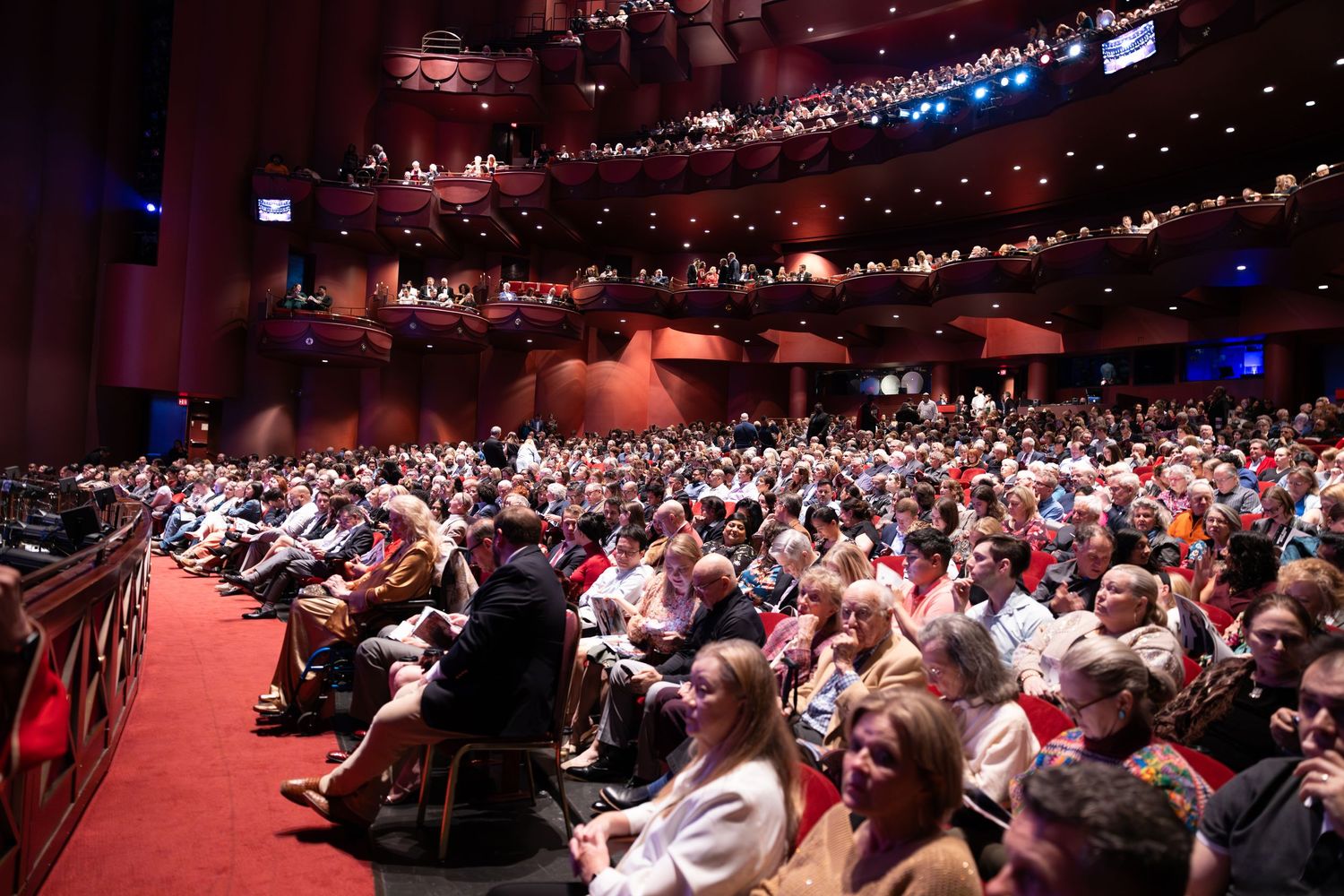 Large audience fills a dimly lit red theater auditorium during a performance at a modern performance venue.