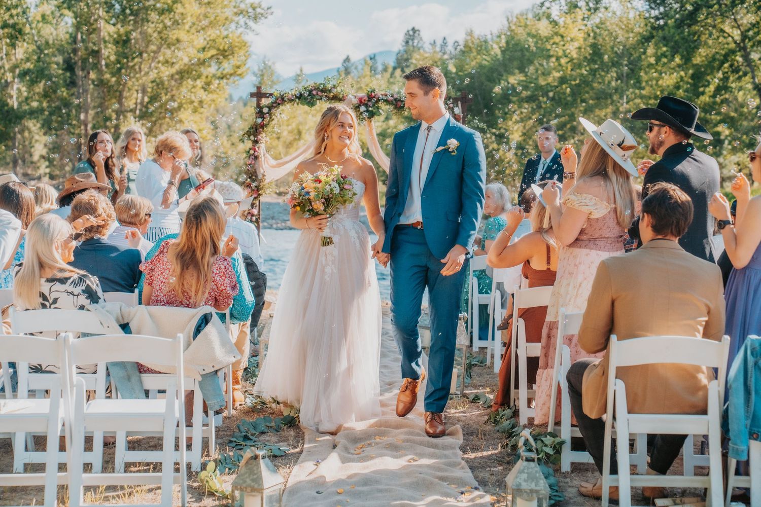 A sunny outdoor wedding ceremony in a wooded setting with white chairs and guests gathered on a stone path.