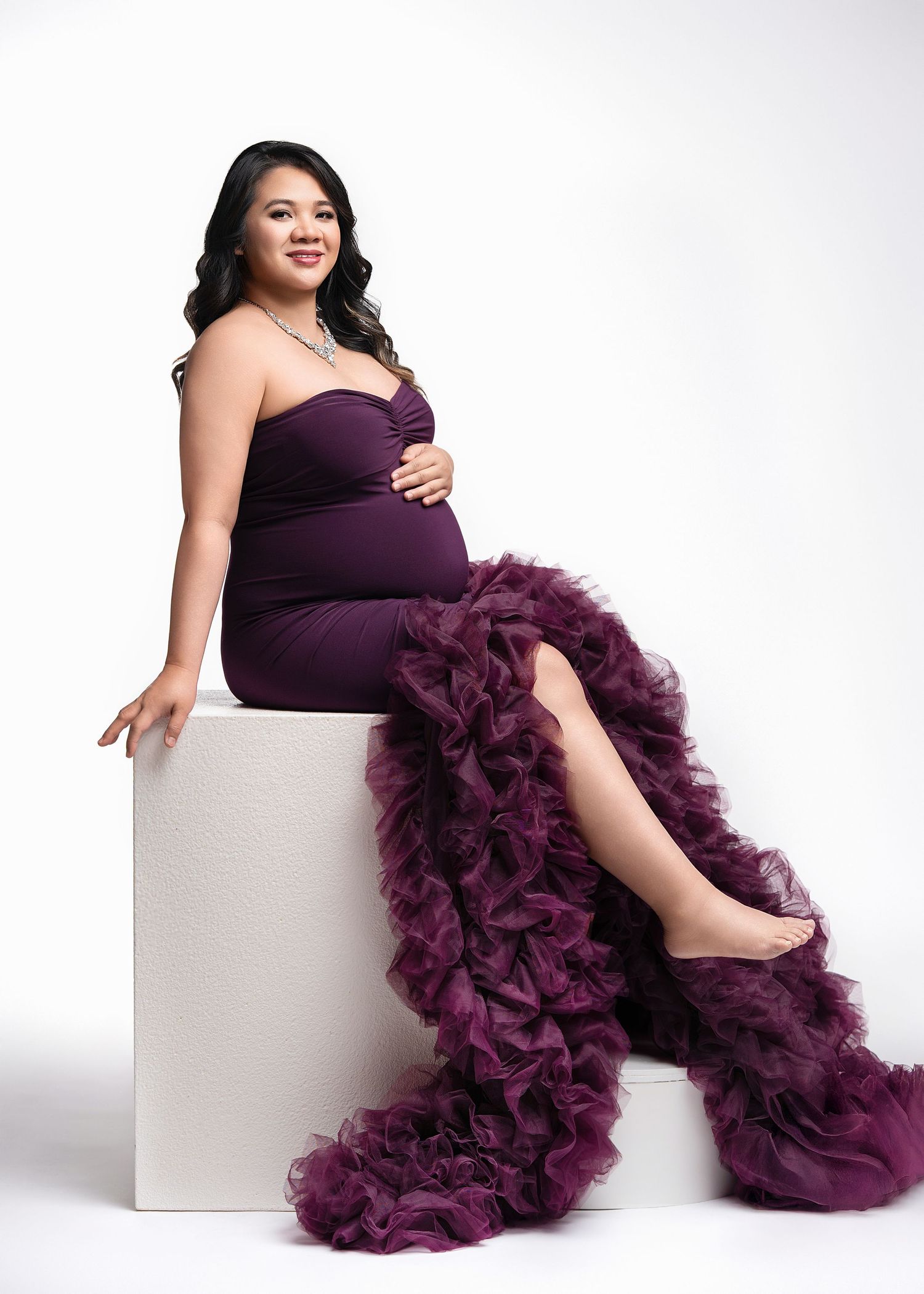 A pregnant woman poses in an elegant purple dress with dramatic ruffled tulle details while sitting on a white cube.