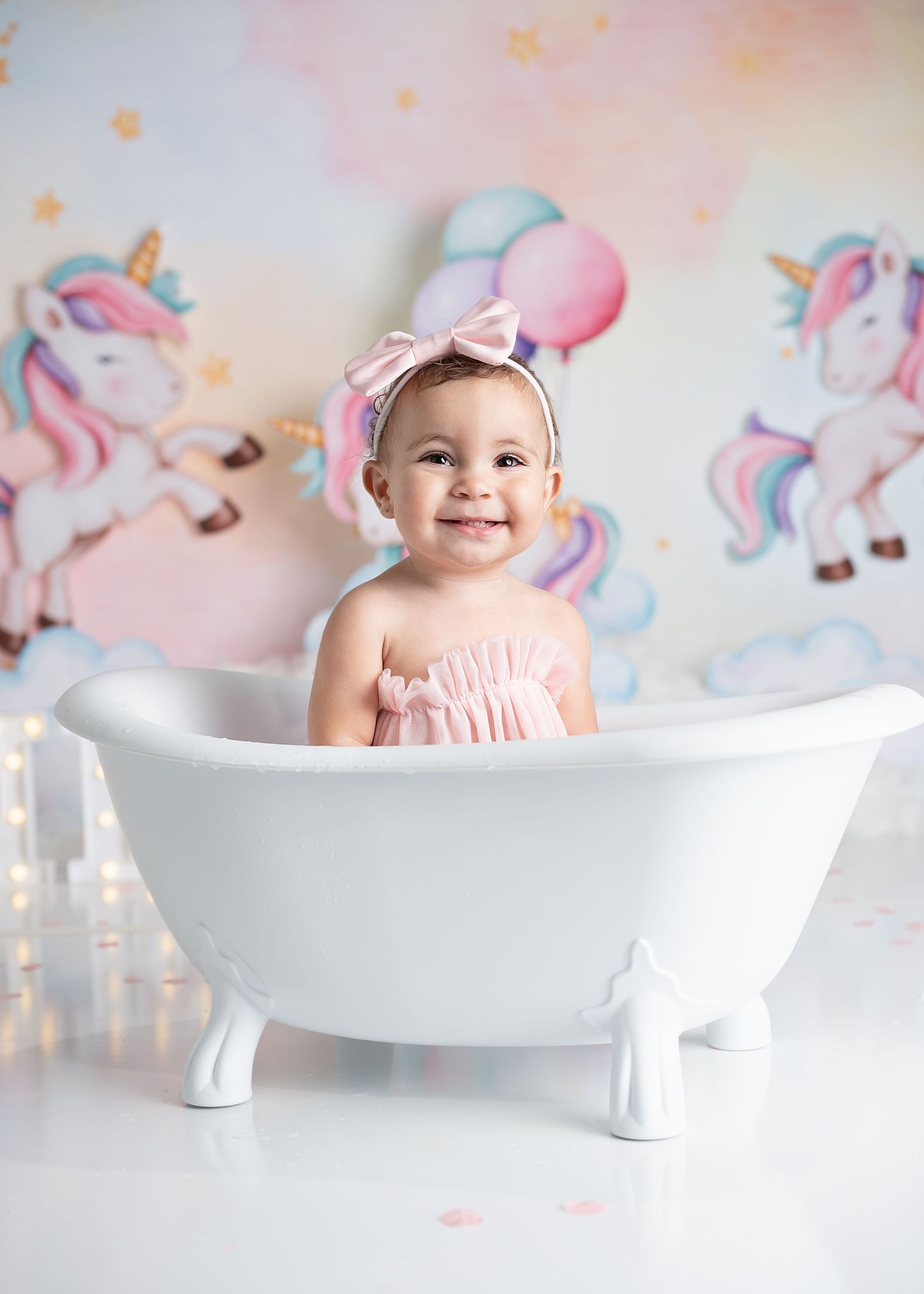 A baby in pink ruffled outfit sits playfully in a white freestanding bathtub against a pastel unicorn-themed backdrop.