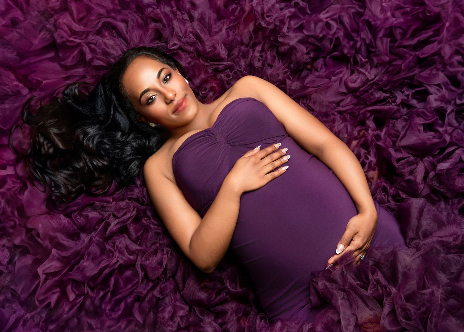 Maternity photo of person in purple dress lying on plush purple fabric background while holding their belly.