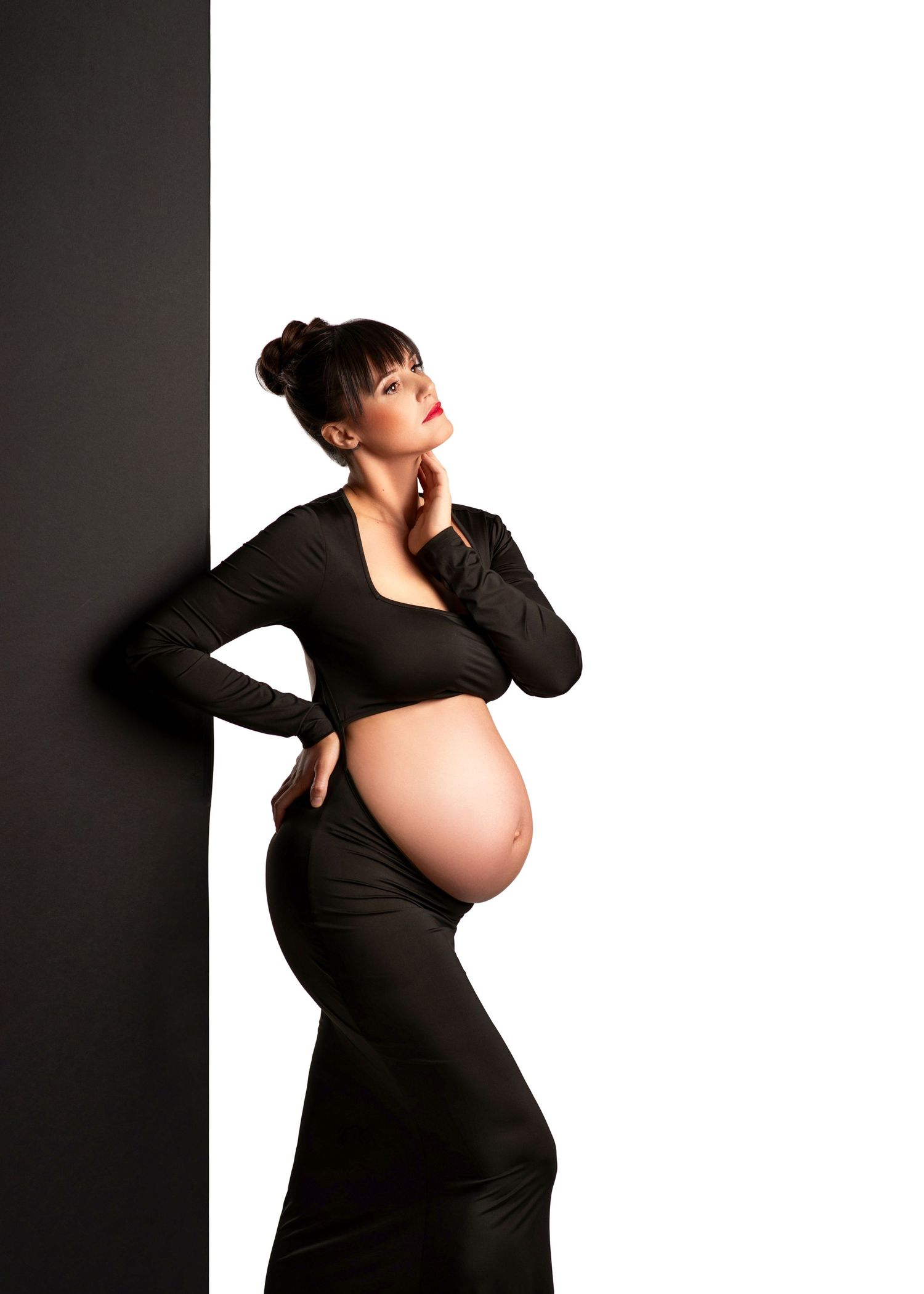 Elegant maternity photo of woman in black outfit posing against dark and light contrasting background.