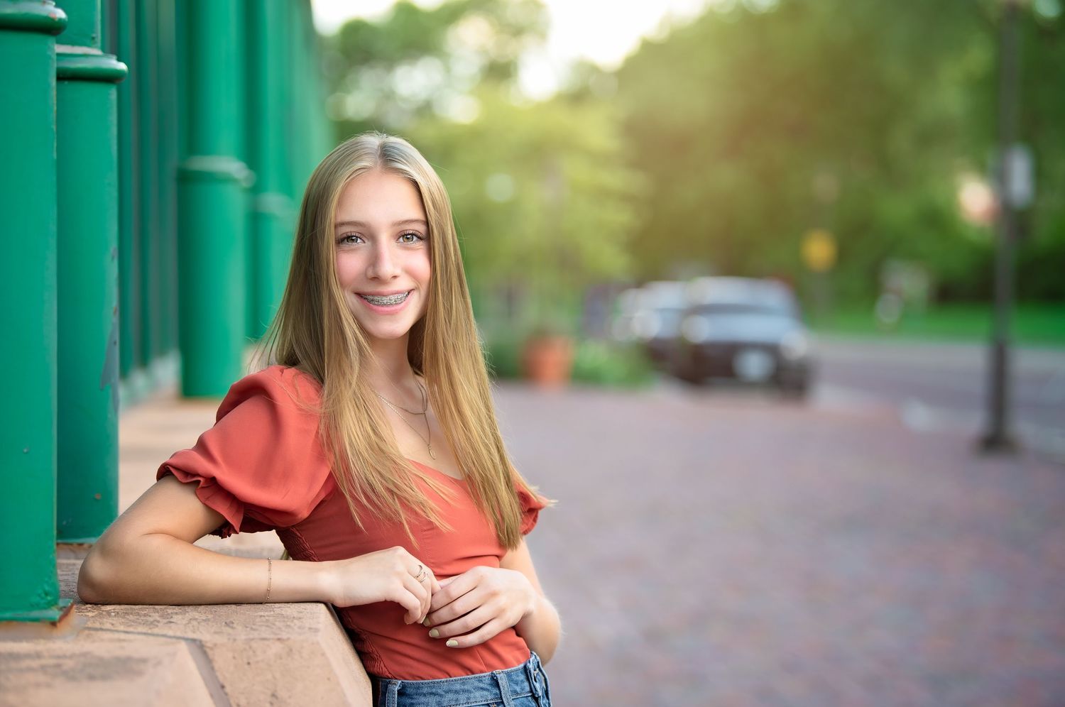 Young woman in coral top leans against green pillar on sunny urban street with soft bokeh background for a minneapolis senior photographer