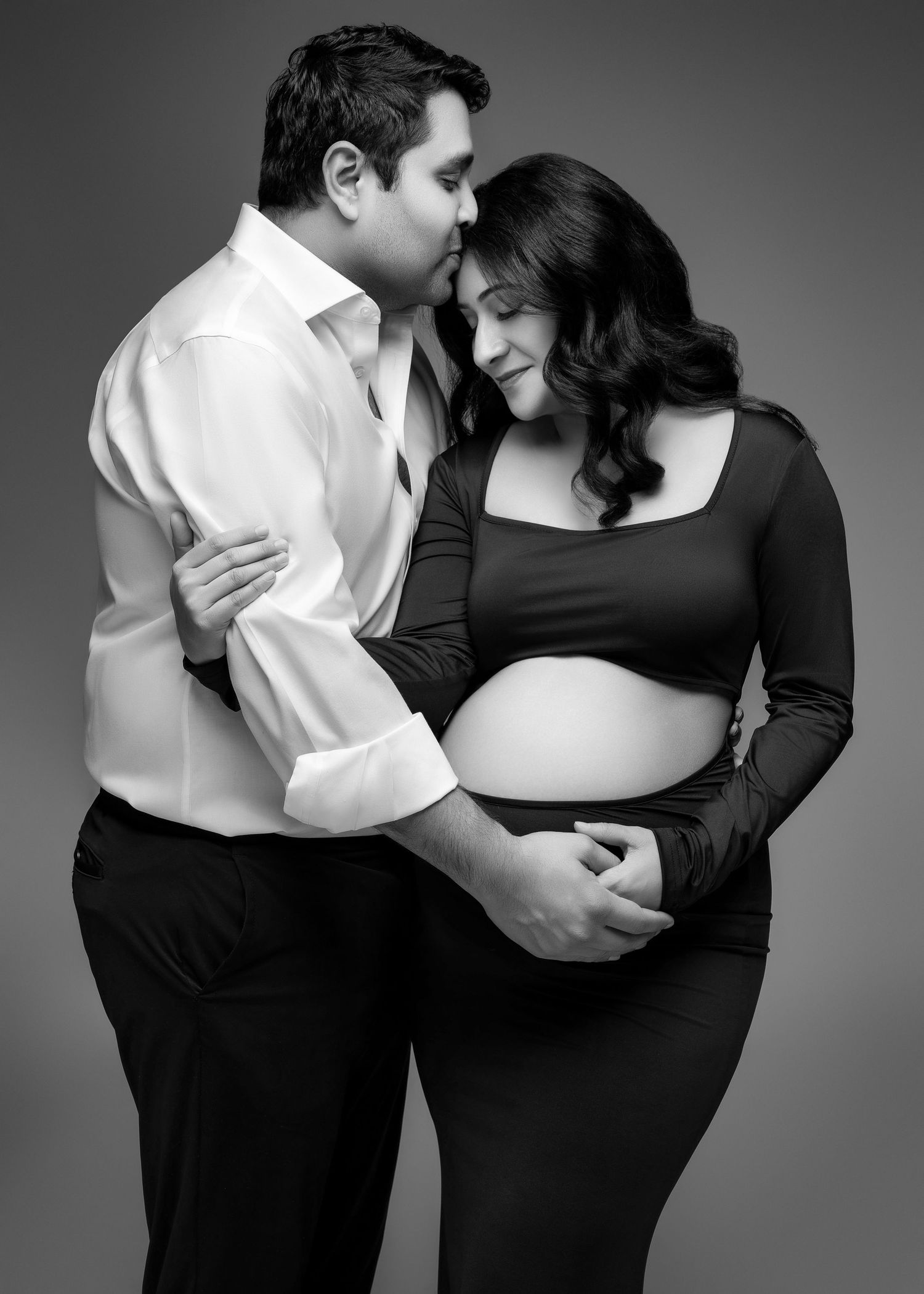 Black and white maternity photo of a couple embracing, with man in white shirt kissing woman in black dress.