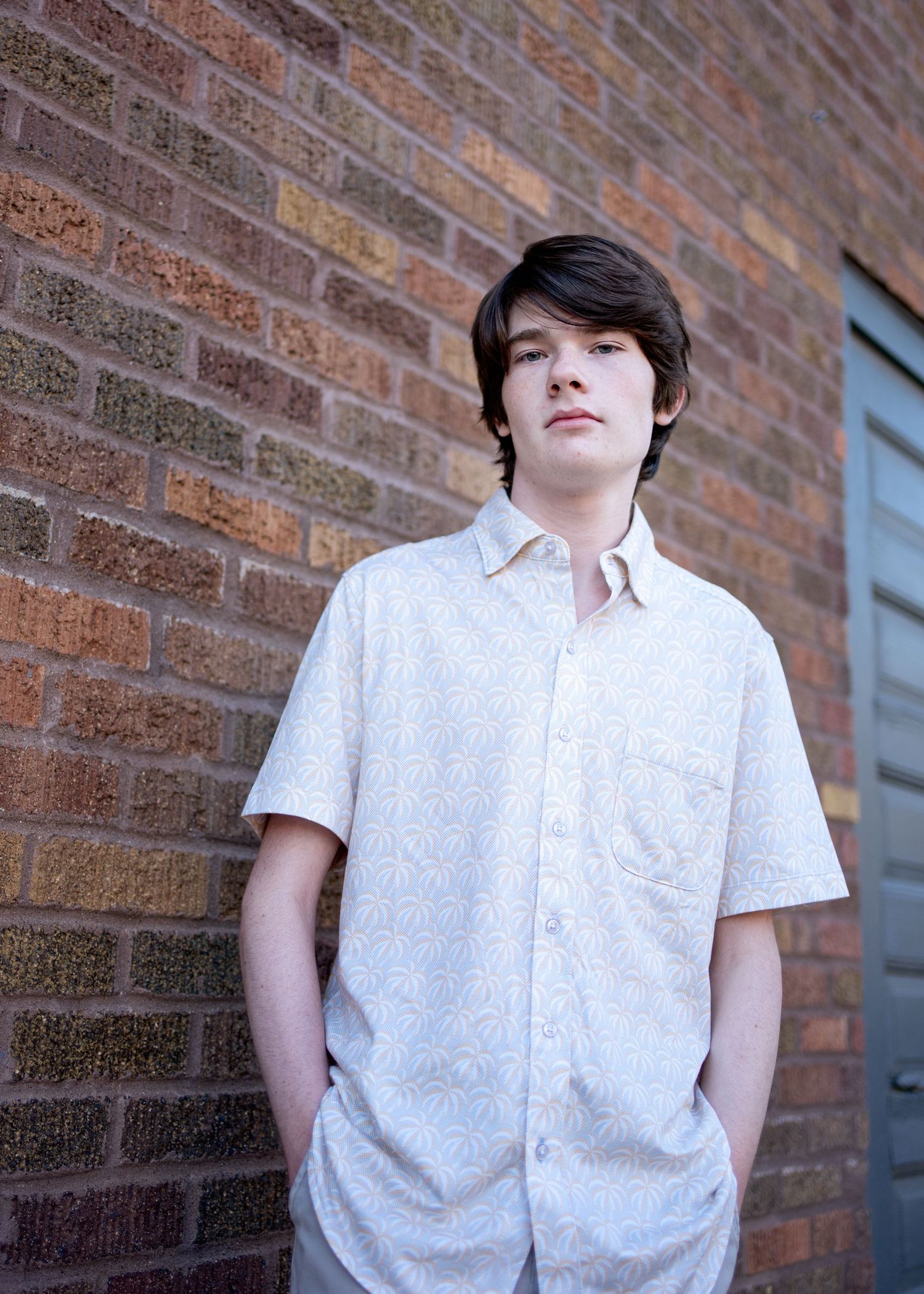 A minneapolis senior in a white short-sleeved button-down shirt stands against a brick wall background in natural lighting.