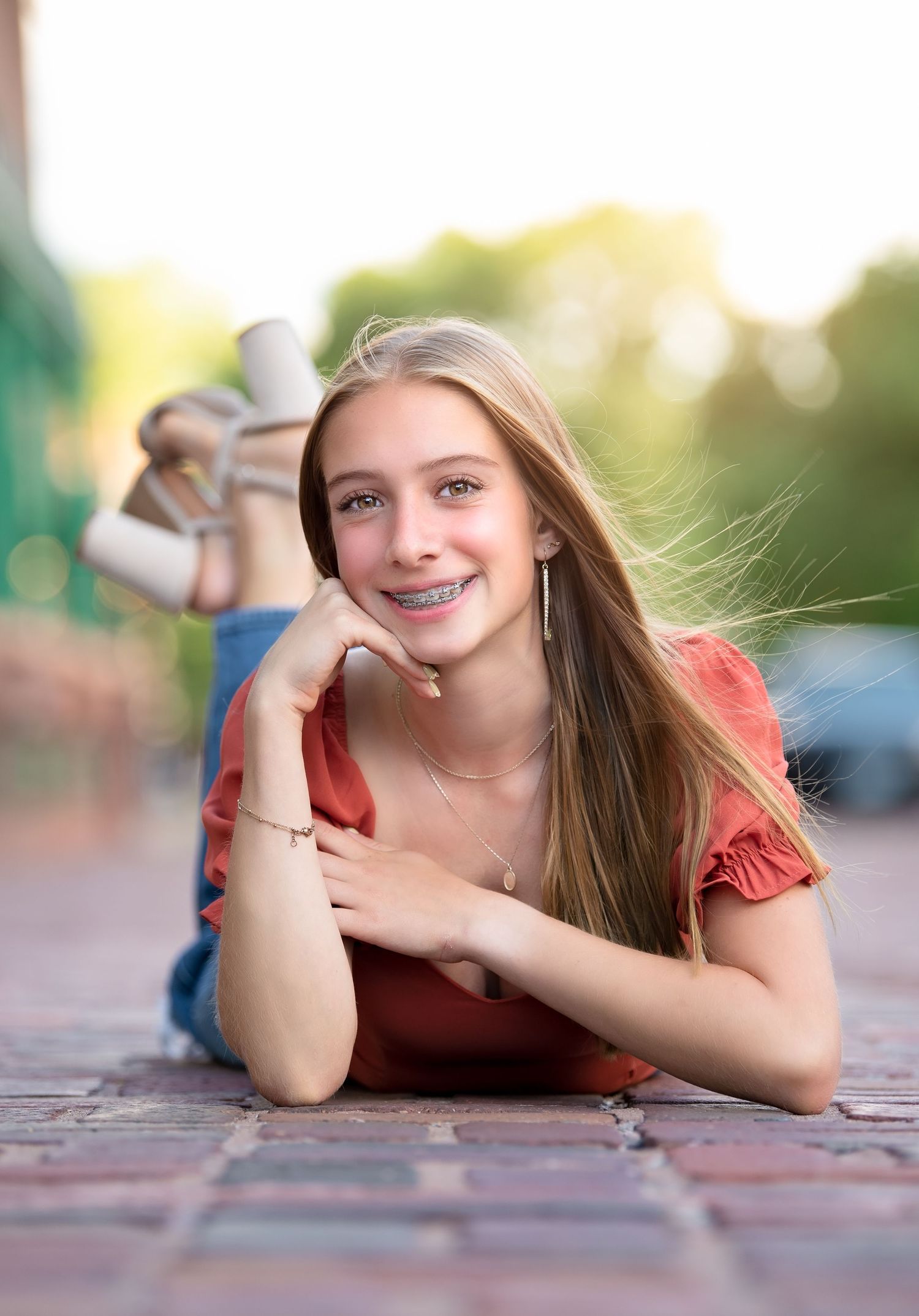 A bright outdoor portrait of someone lying on brick pavement in a red top with long hair and braces smiling at the camera for senior photos