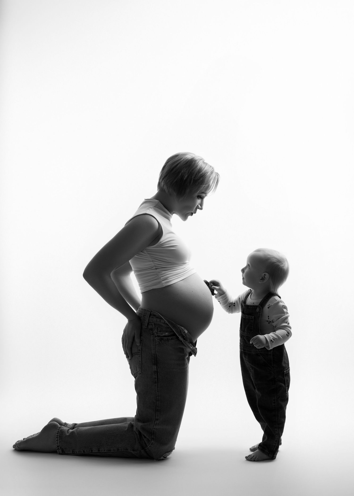 a pregnant woman kneels next to her child who drives a car across her belly