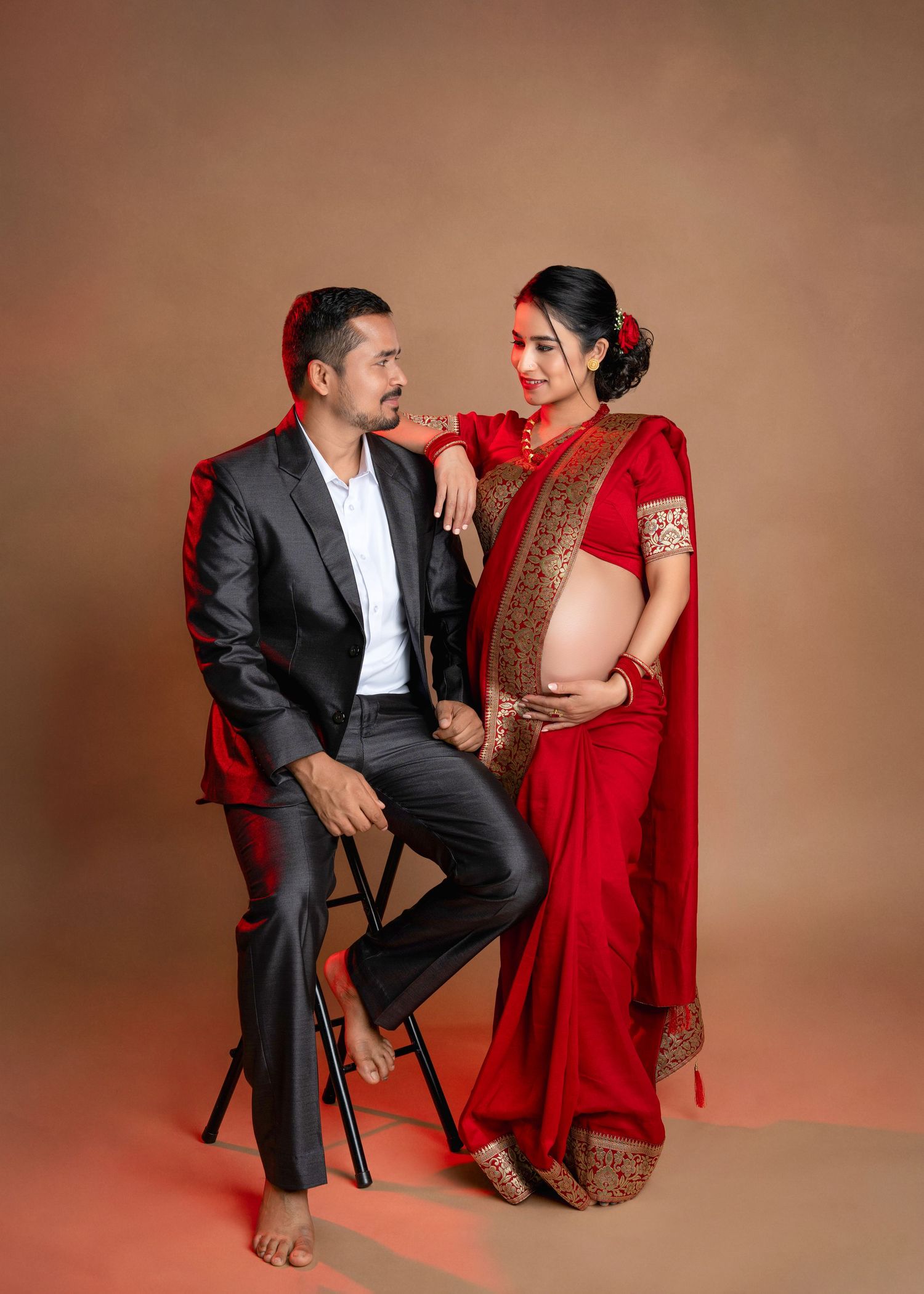 Studio portrait of a couple in formal attire, with one person in a suit seated on a stool and another in a red saree standing beside.