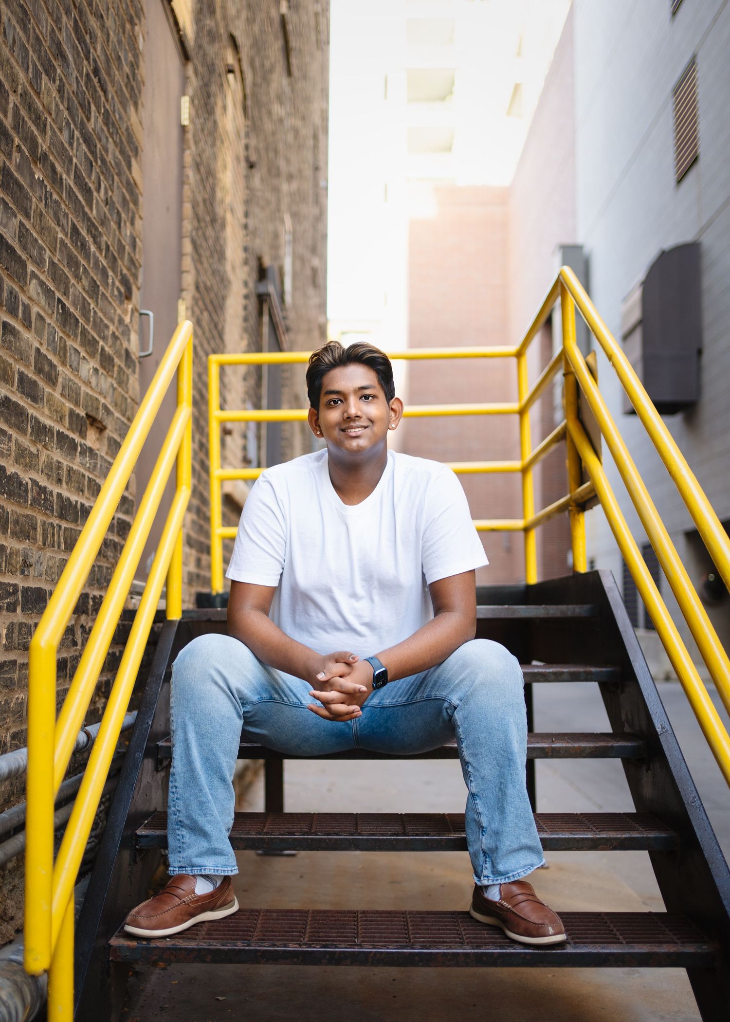 minneapolis senior photography of a teen in a white t-shirt and jeans sitting on stairs with bright yellow handrails between brick walls.
