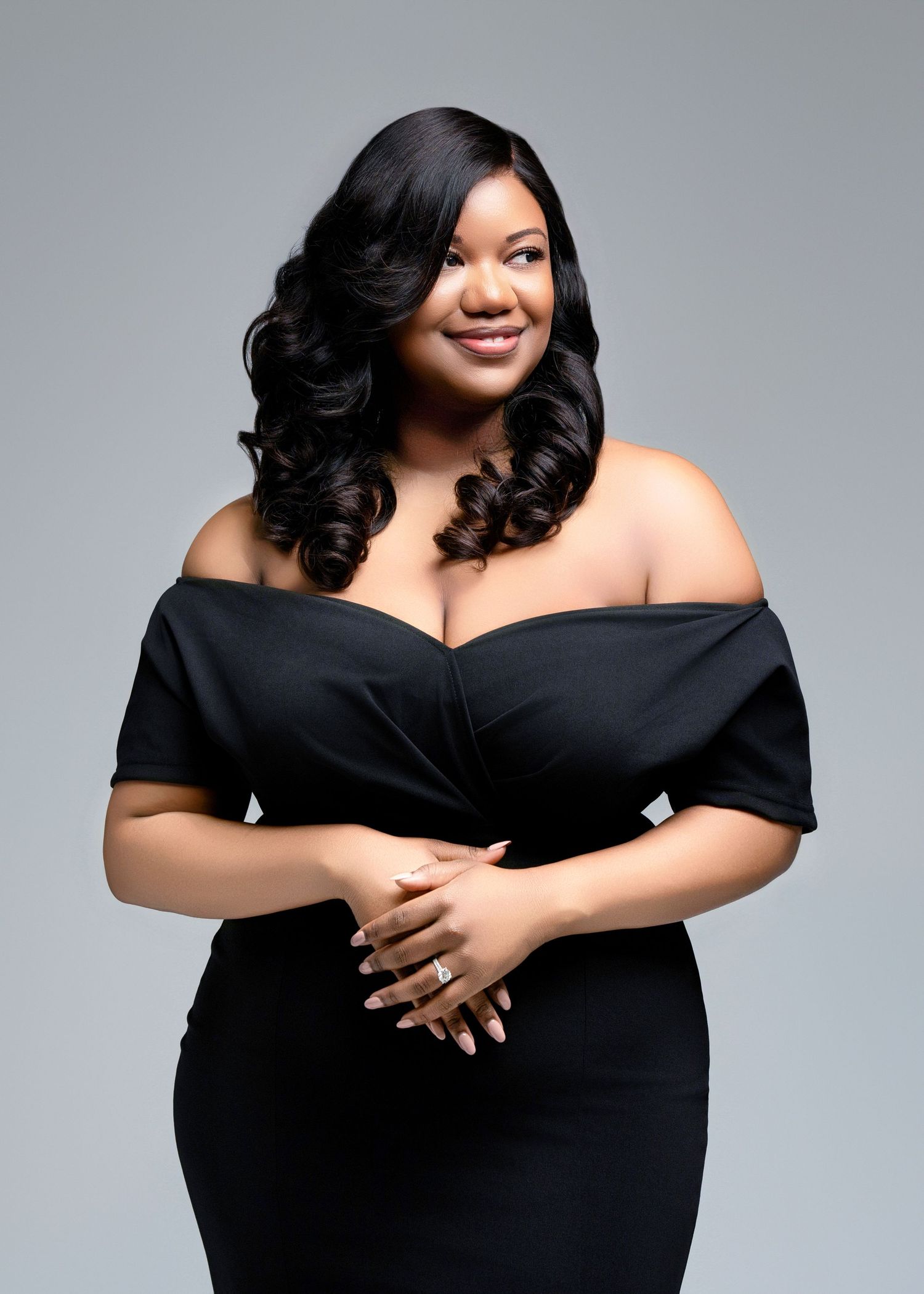 A woman in an elegant black off-shoulder dress poses confidently against a gray background with styled curly hair.
