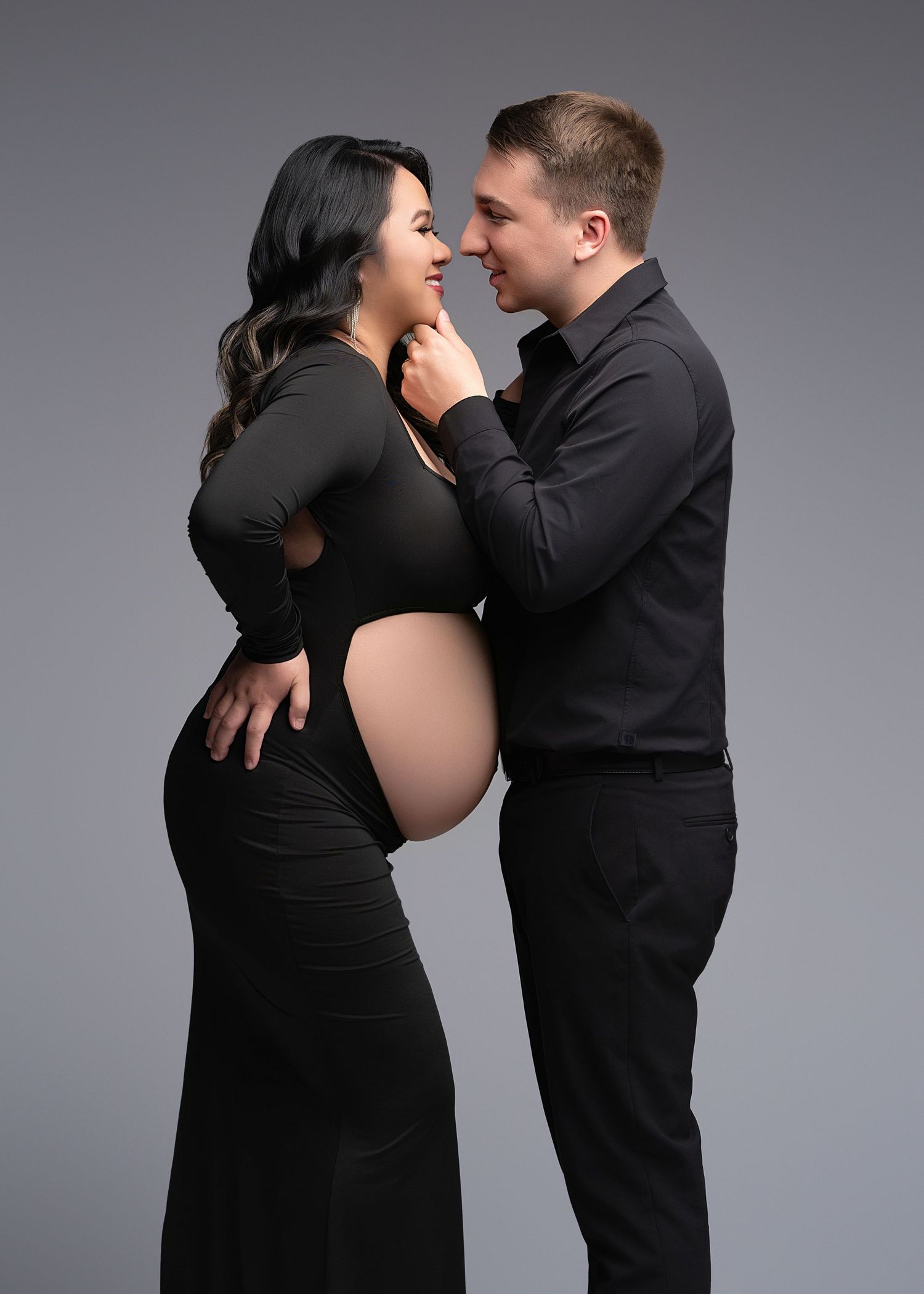 An expecting couple dressed in black embrace intimately during a maternity photoshoot against a gray backdrop.