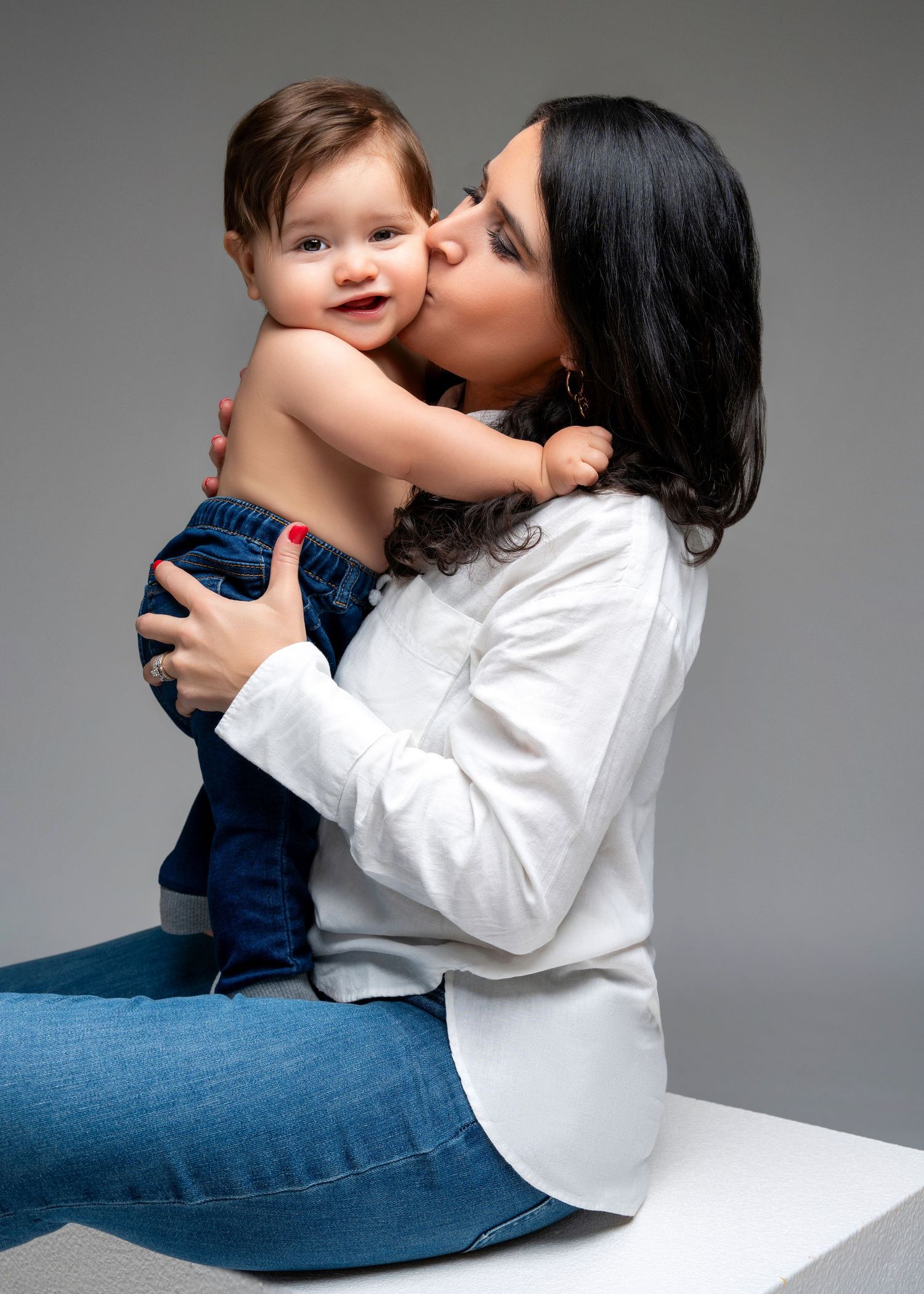 a mother sits on a white cube while pressing a kiss to a her child's cheek