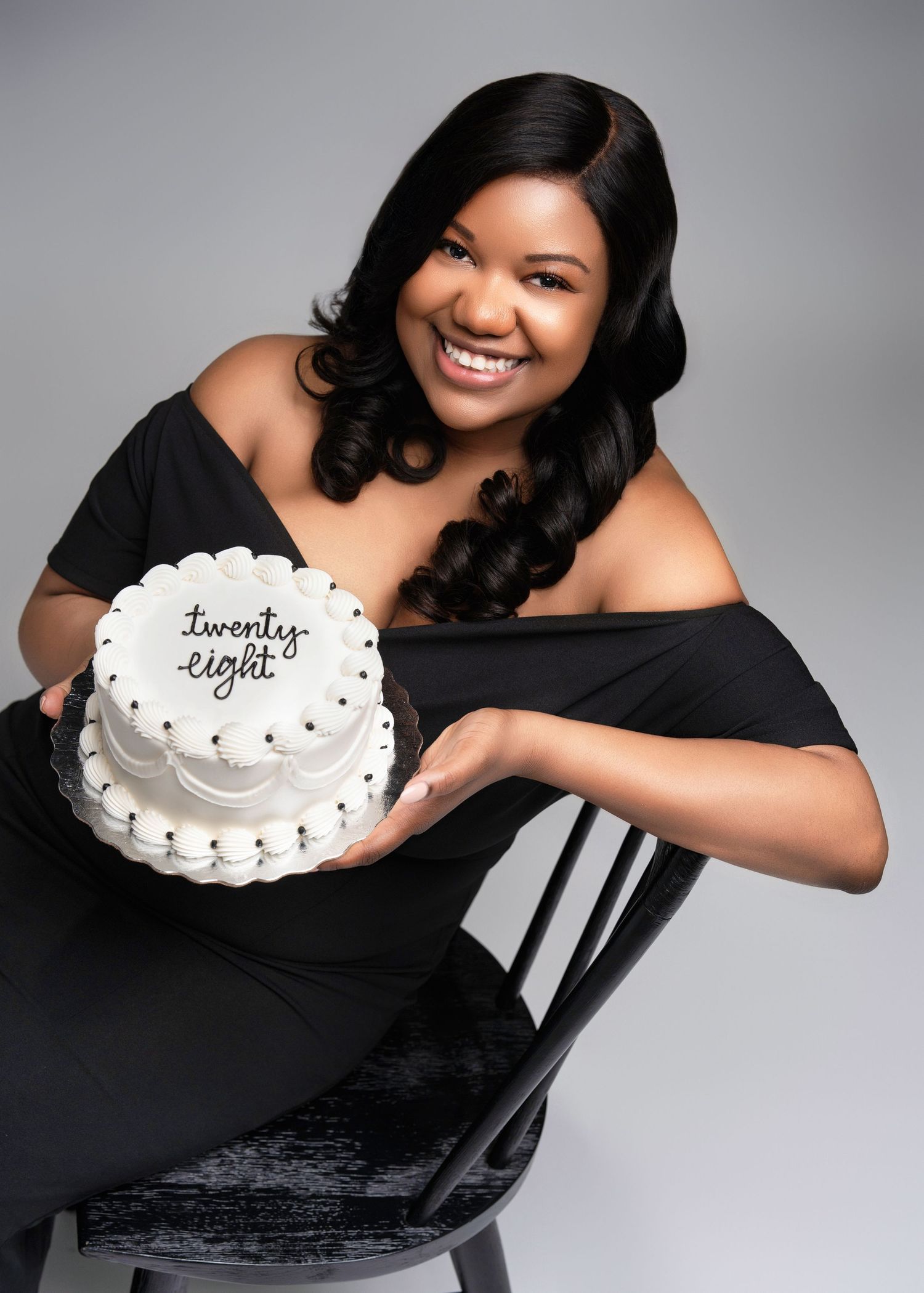 A woman in a black off-shoulder dress smiles while holding a white birthday cake with 