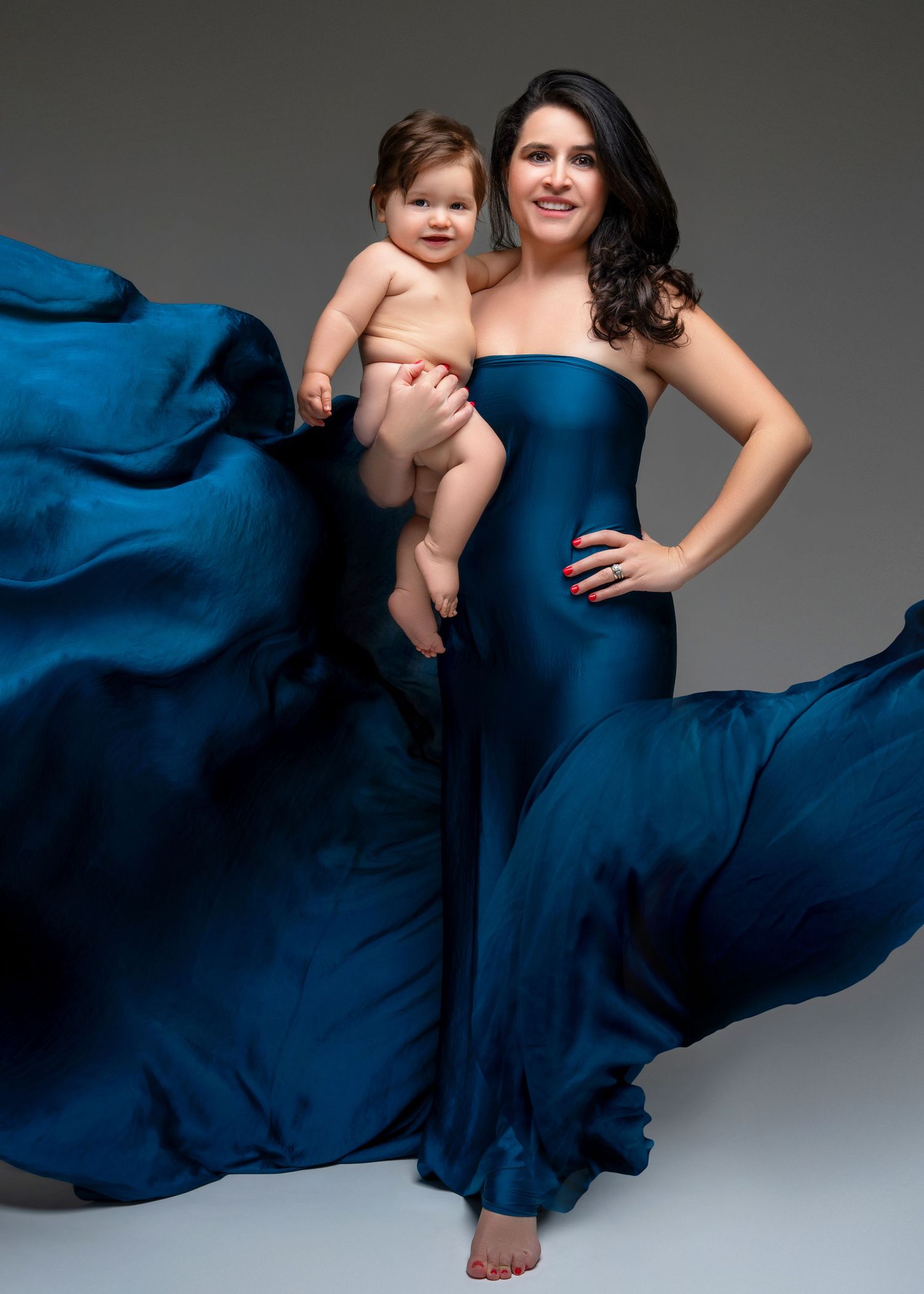 a woman wearing a flowing blue gown stands with her child in an arm