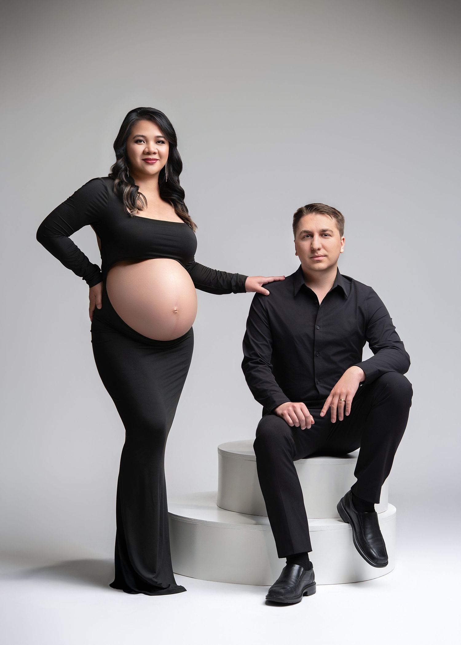 Elegant maternity photo of a couple in black attire posing against a light grey studio background.