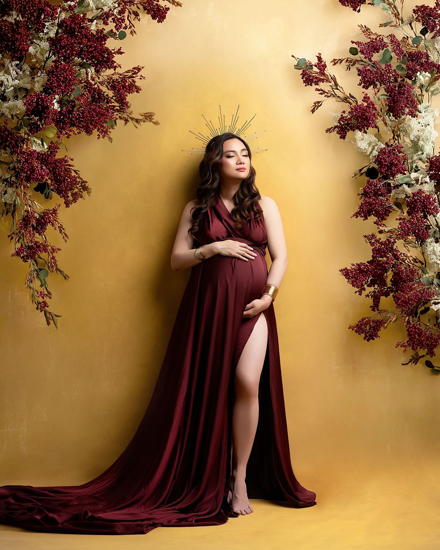 Artistic maternity portrait in Minneapolis against warm yellow backdrop with burgundy flowers and flowing wine-colored fabric draping