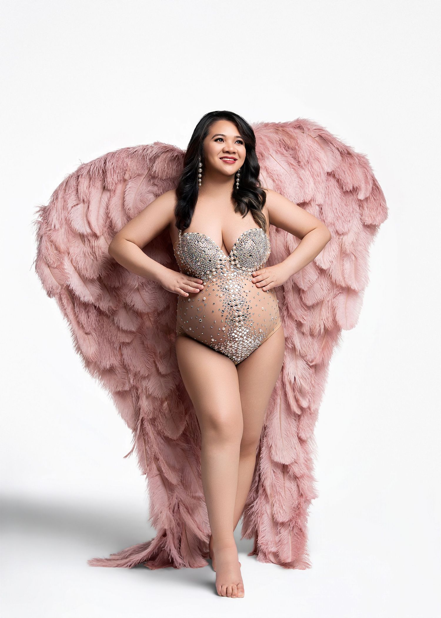 A performer in a bedazzled bodysuit poses confidently against a pink feathered wing backdrop on white background.