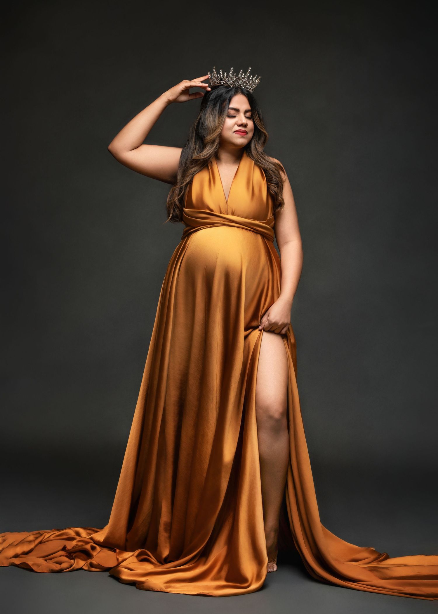 a woman wearing a flowing golden gown rests a hand on her crown