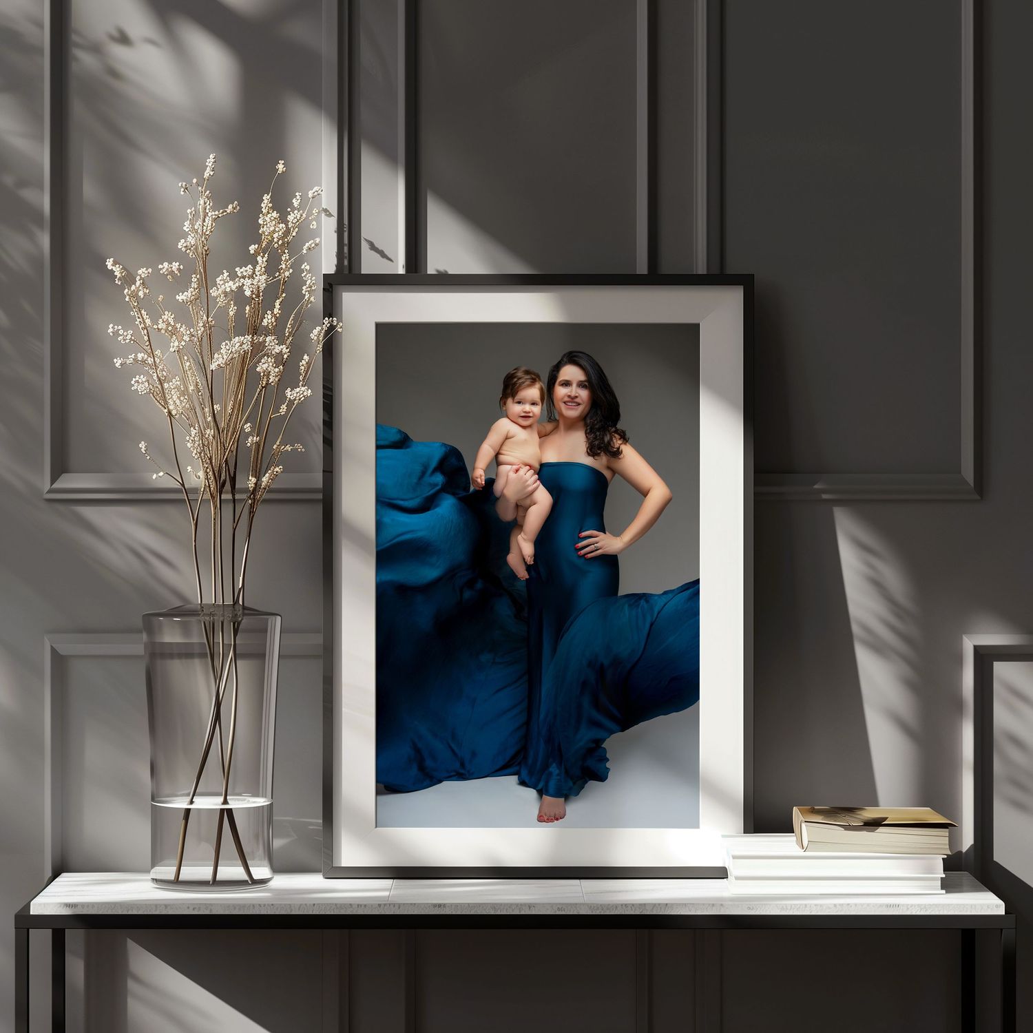 Framed photograph displayed on white shelf with dried flowers in vase and natural shadows on gray wall.