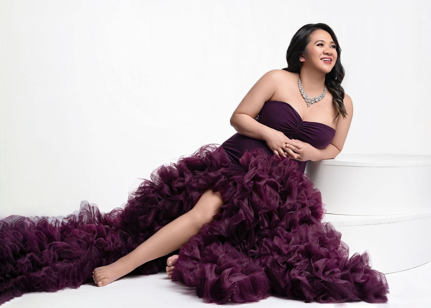 A glamorous figure poses in a flowing plum-colored tulle gown against a white studio backdrop.