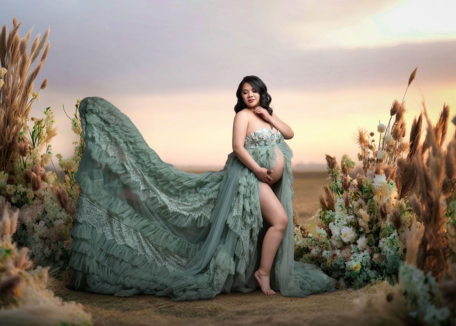 An ethereal flowy green maternity gown billows in the breeze amidst dried flowers at sunset.