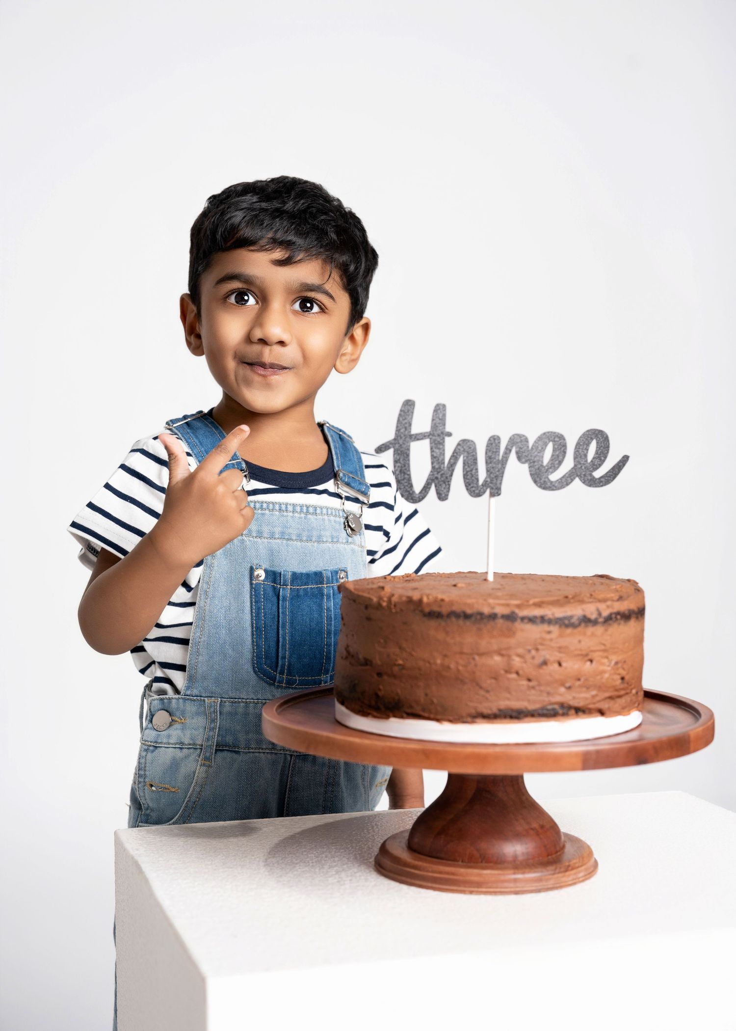 a young boy brings his finger towards his mouth after touching the frosting on his three year birthday cake