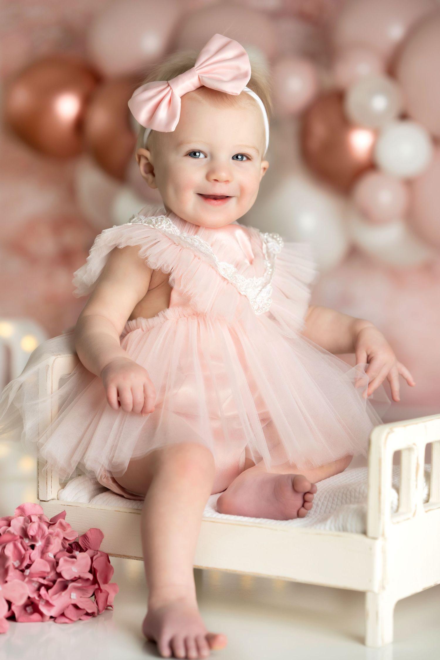 Baby wearing pink tulle dress and bow sitting on white bench with rose gold balloons in background.