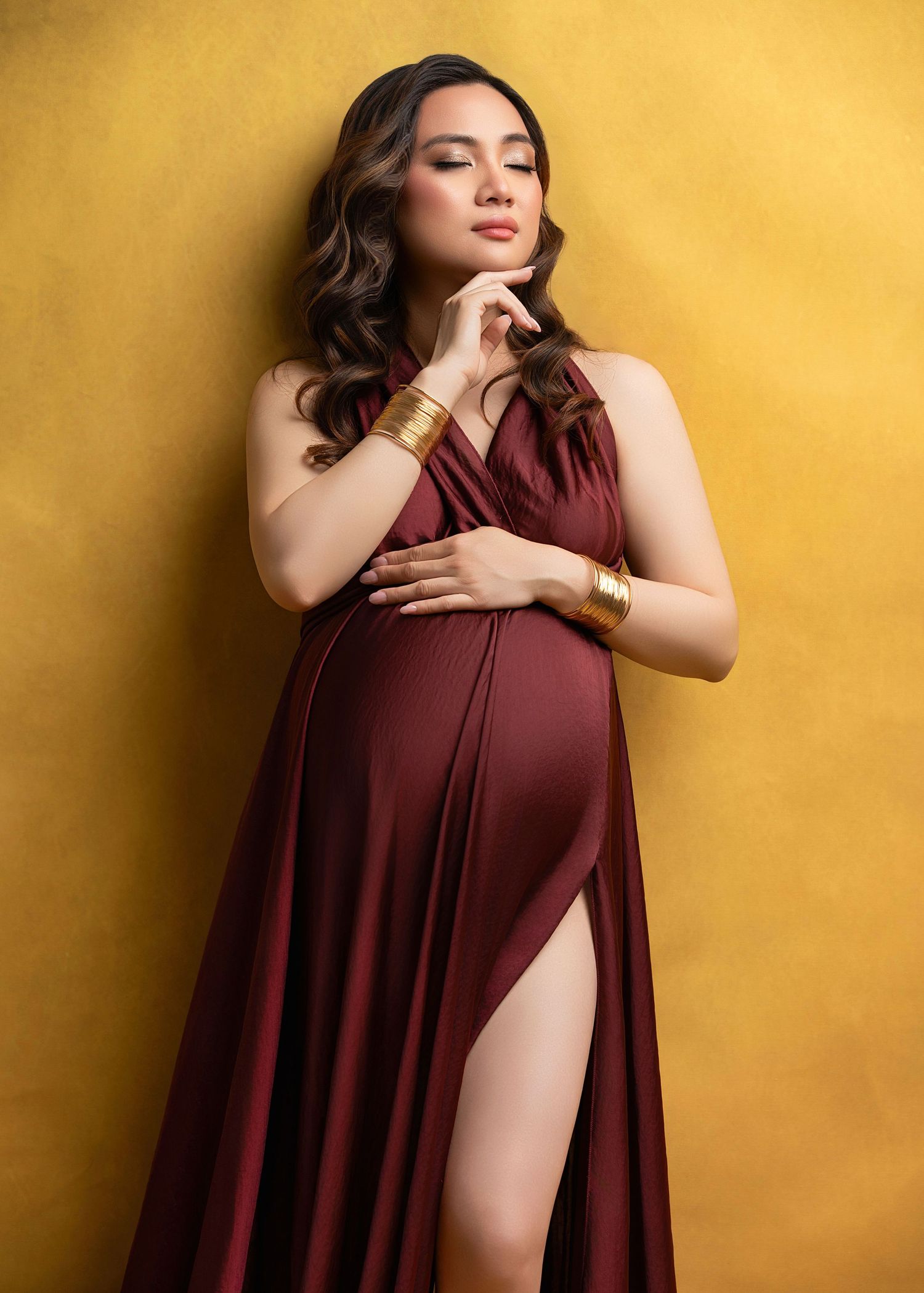 A pregnant woman in a burgundy formal dress poses elegantly against a golden yellow backdrop.