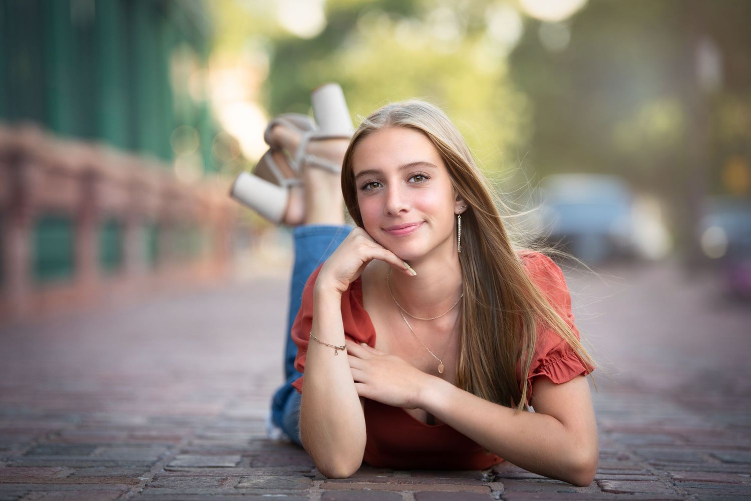 A young person lies on a brick sidewalk with long straight hair and a red shirt, photographed with a soft blurred background.