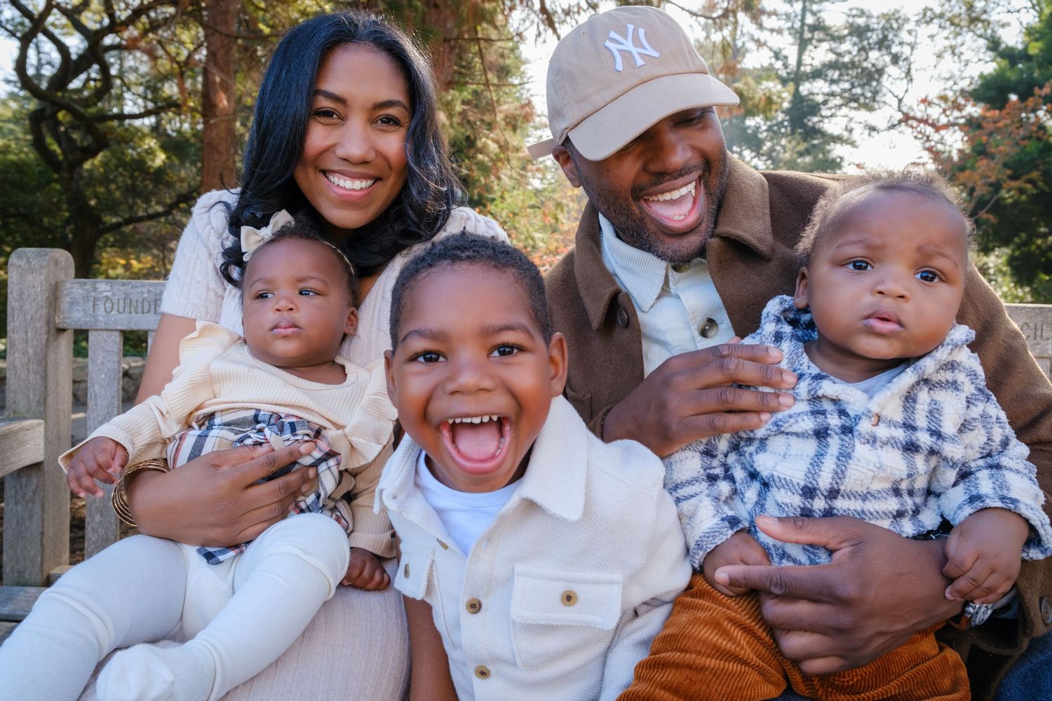 A joyful family sitting together on a wooden bench outdoors during autumn, with two young children in coordinated outfits.