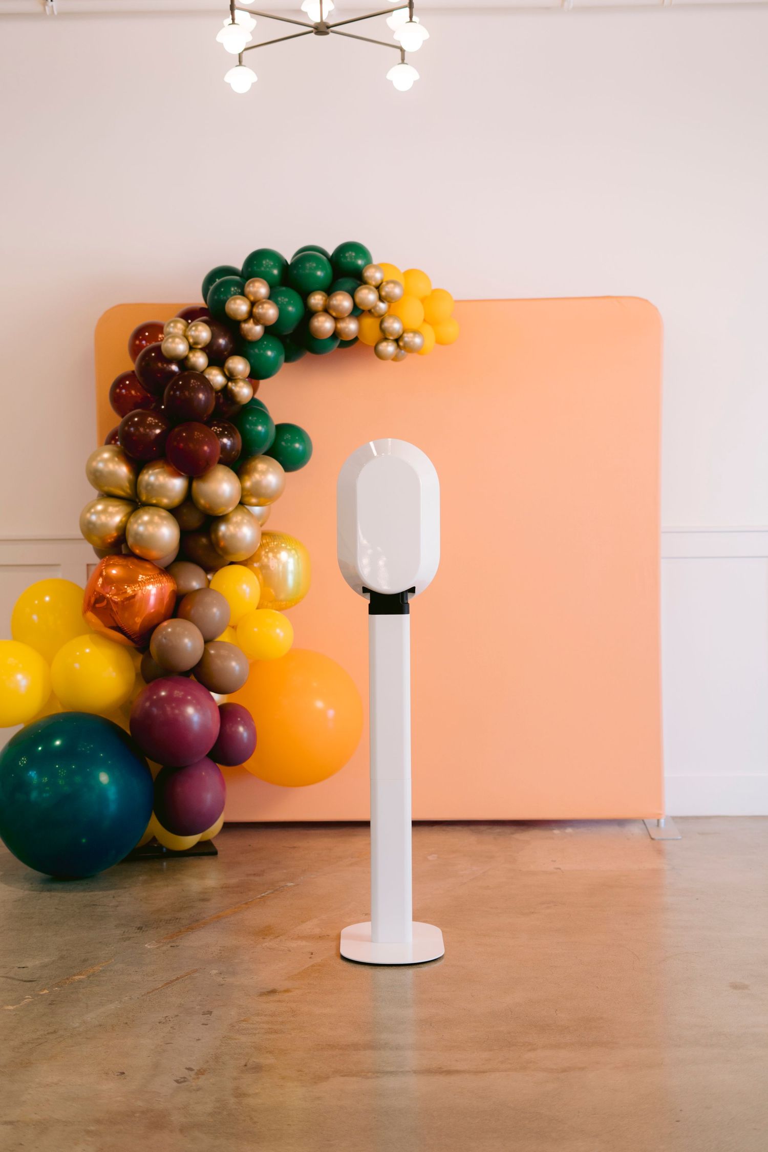 A modern white cylindrical robot stands on a wooden floor next to a colorful balloon arch installation in orange lighting.