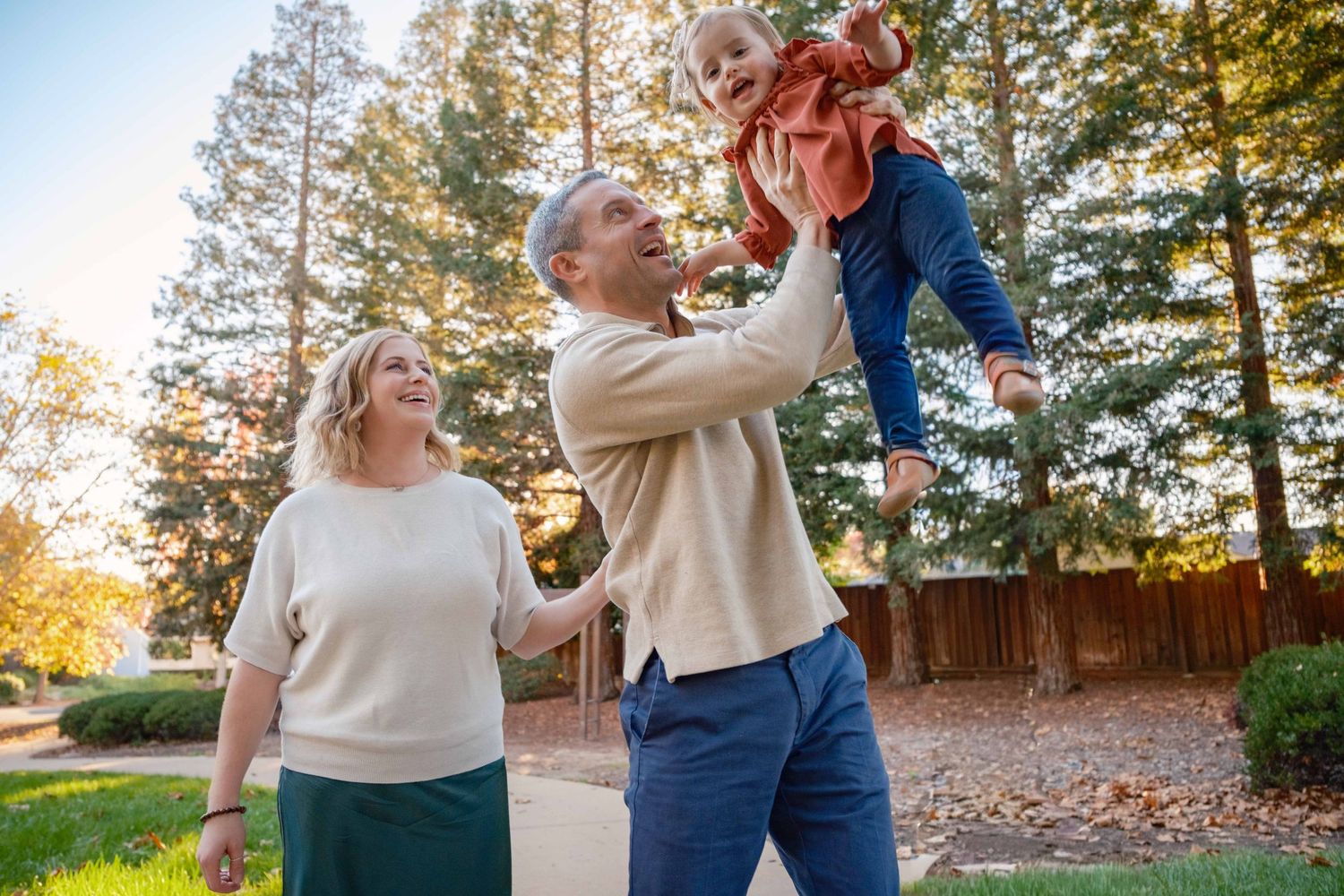 A couple embraces outdoors during golden hour in a backyard with tall trees and autumn leaves.